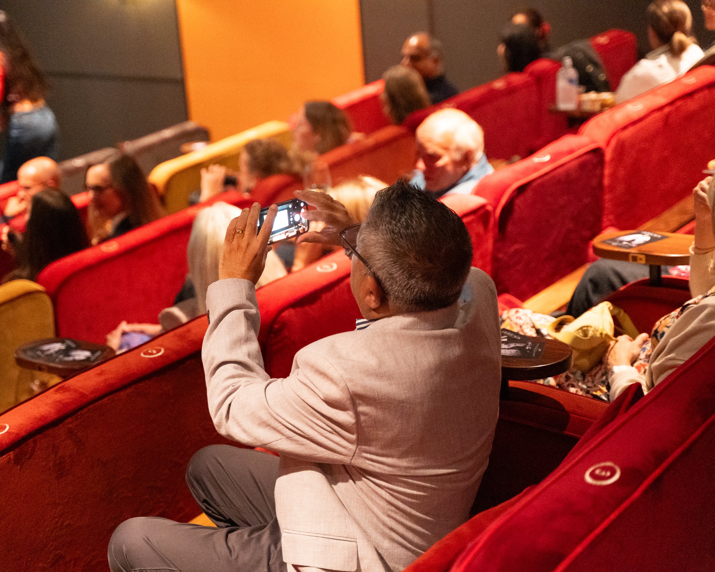 People seated in red theater seats, with some taking photos or videos, in a dimly lit auditorium.