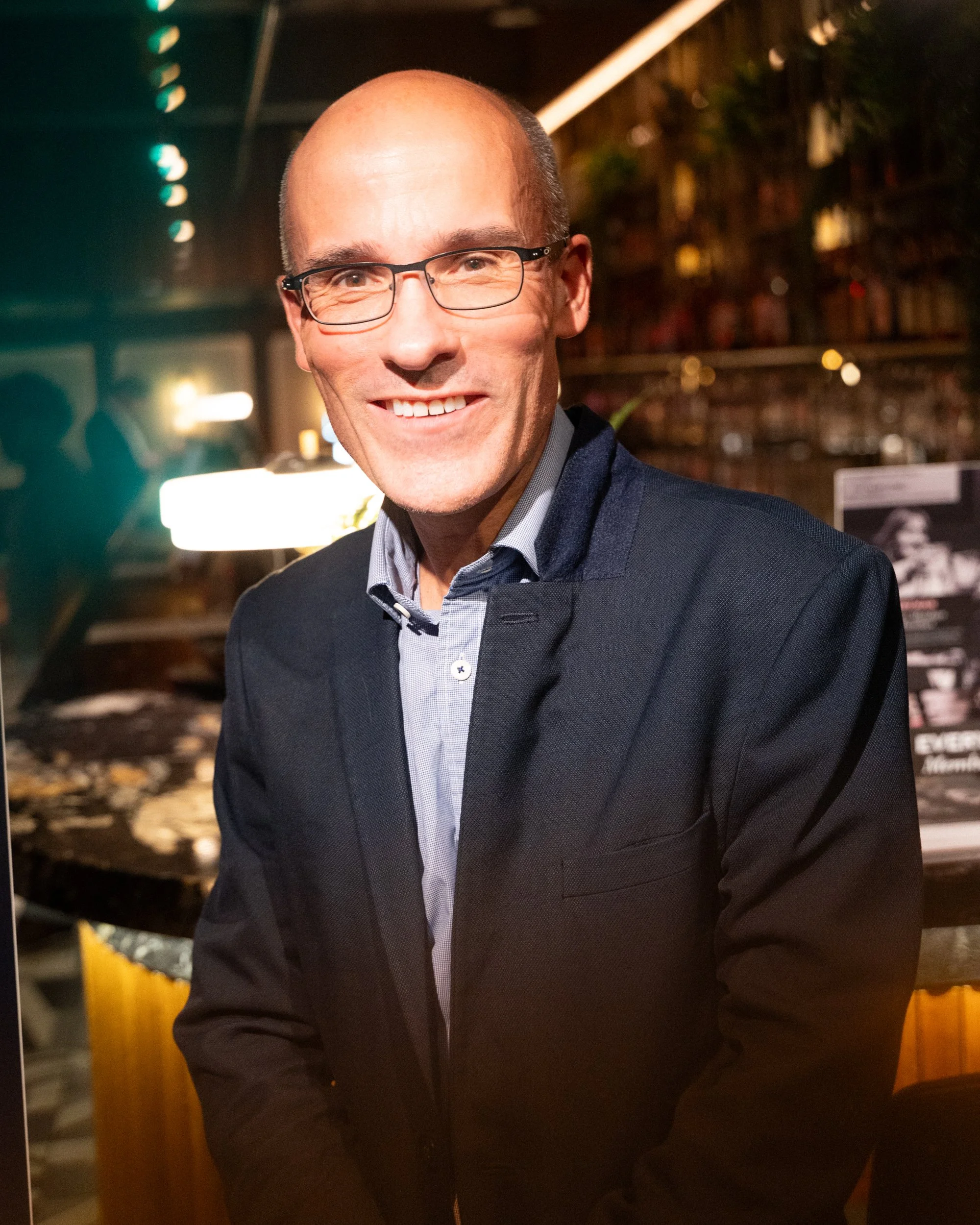 A smiling man with glasses in a dark indoor setting, wearing a dark suit and light collared shirt.