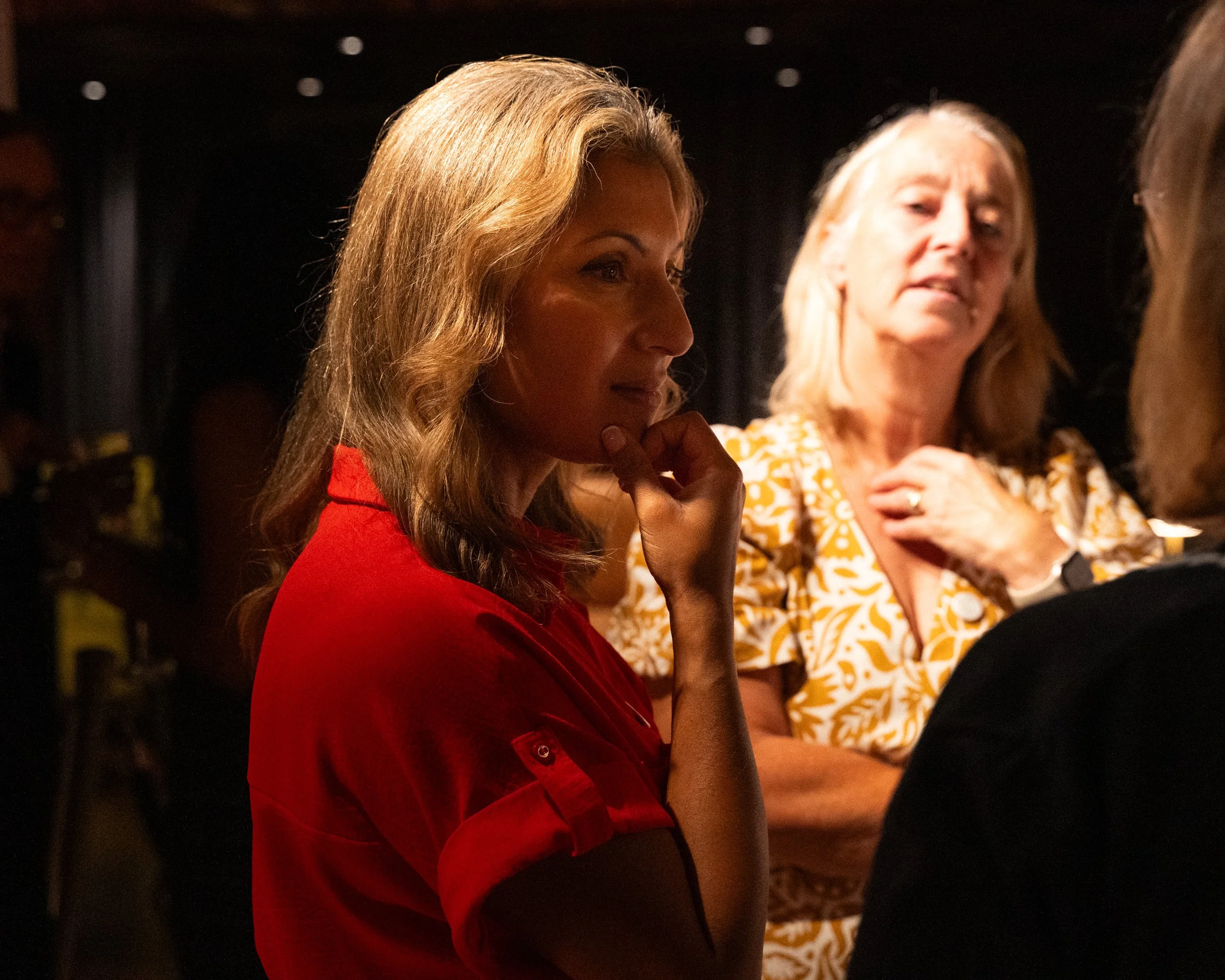 Two women sitting at a table, one with long, wavy blonde hair wearing a red top, and the other with blonde hair wearing a yellow patterned dress, both appearing to be in deep thought or conversation in a dimly lit setting.