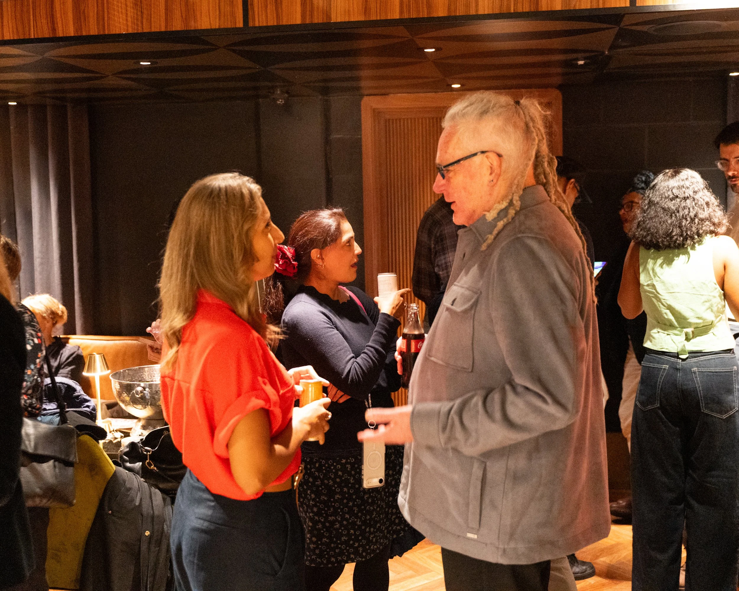 People socializing at an indoor event with wooden walls and warm lighting, including two women and a man engaged in conversation.