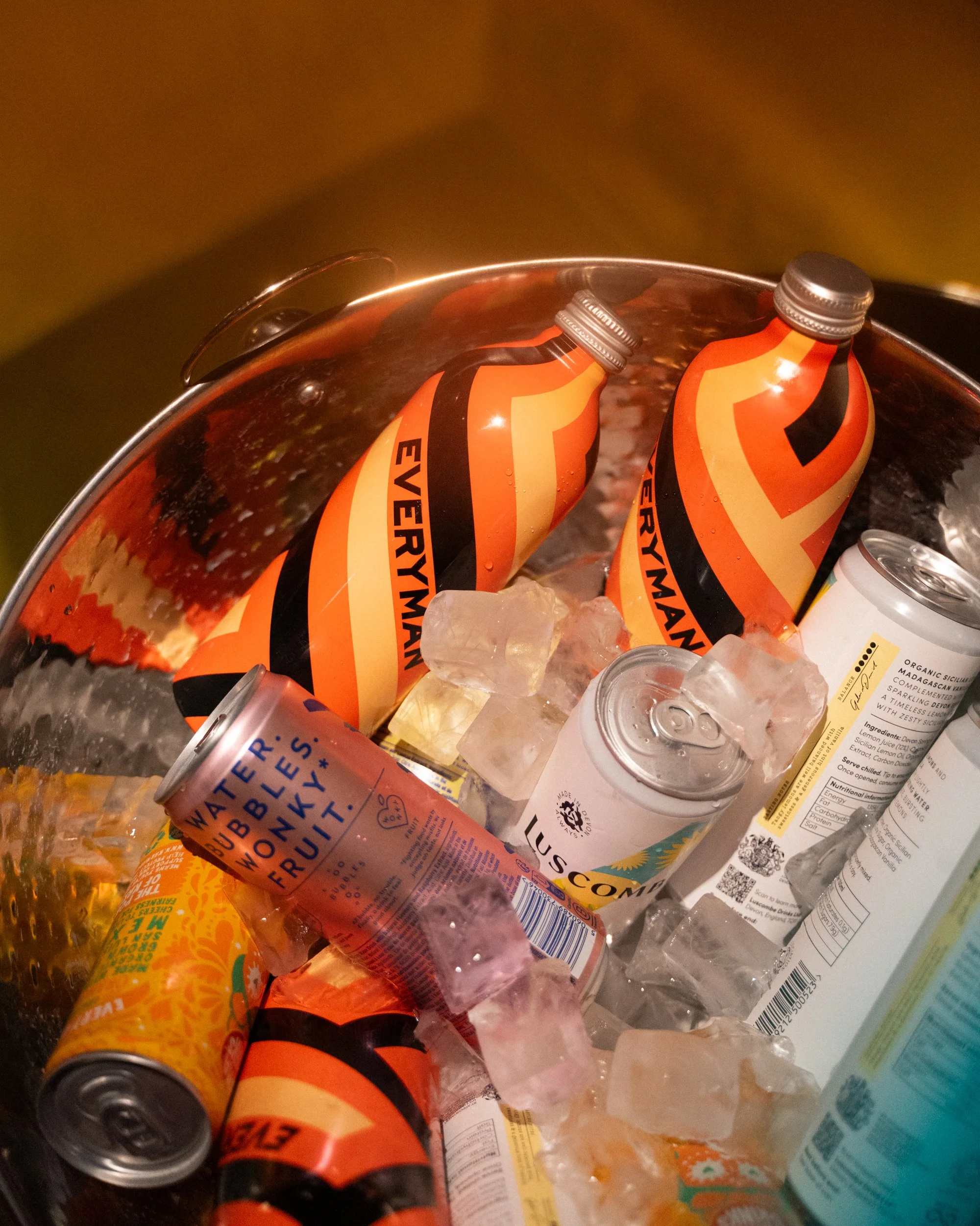 Two orange and black cans of energy drink in a bucket of ice, surrounded by various juice and beverage bottles.