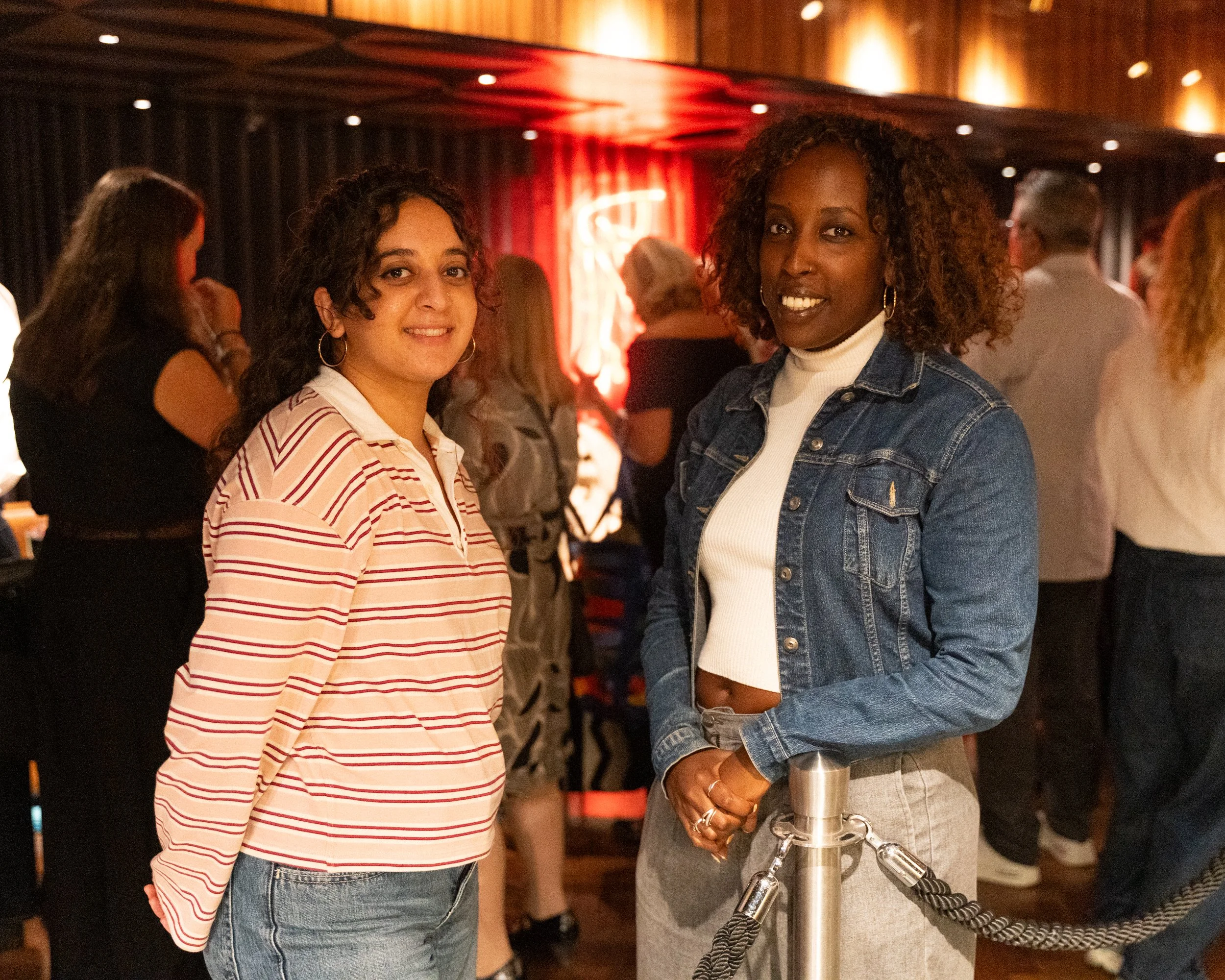 Two women standing in a cozy indoor setting, smiling at the camera, with other people in the background, warm lighting, and wood-paneled walls.