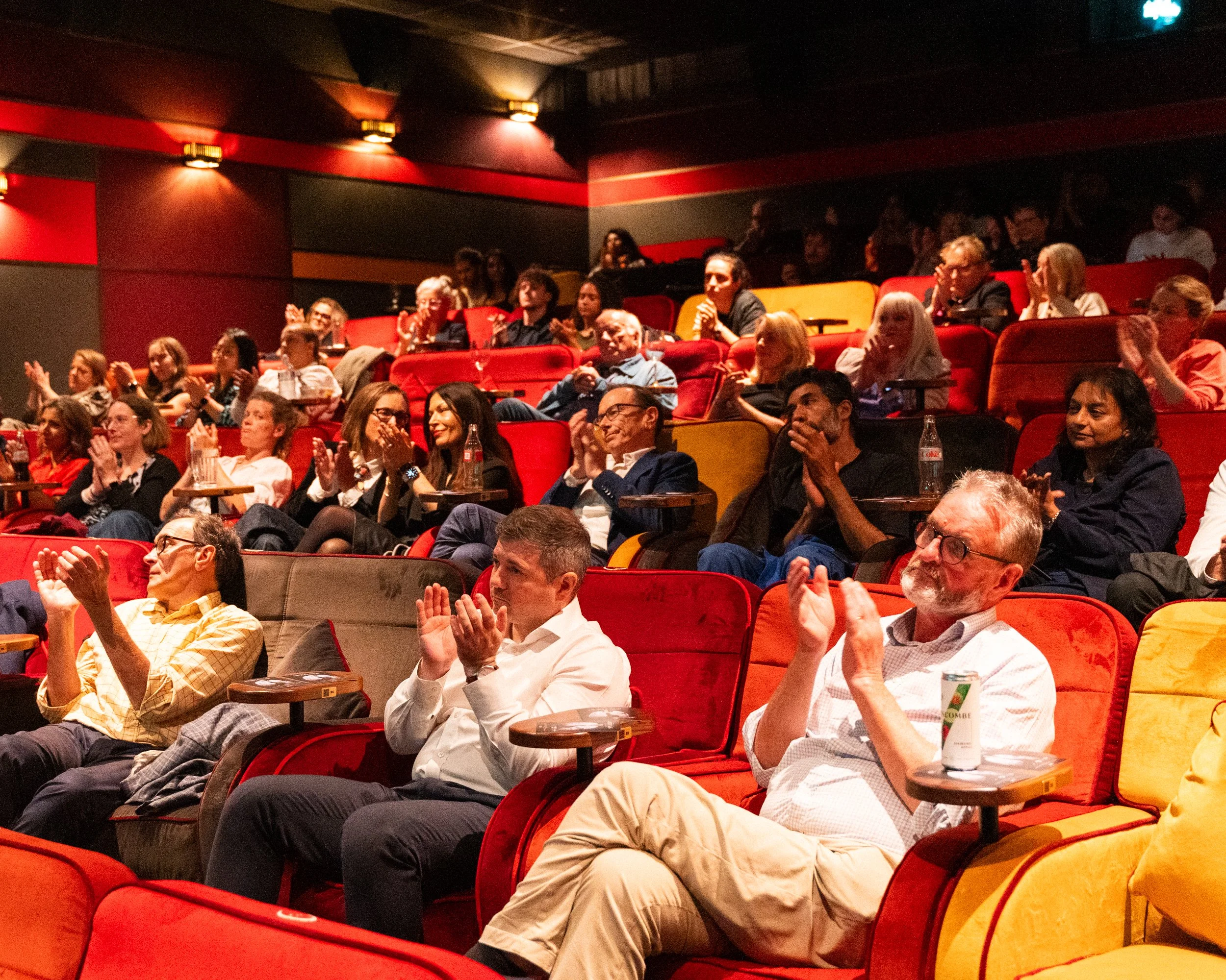 Crowd of people sitting in red theater seats, watching and clapping during a performance, with some drinking beverages, in a dimly lit auditorium.