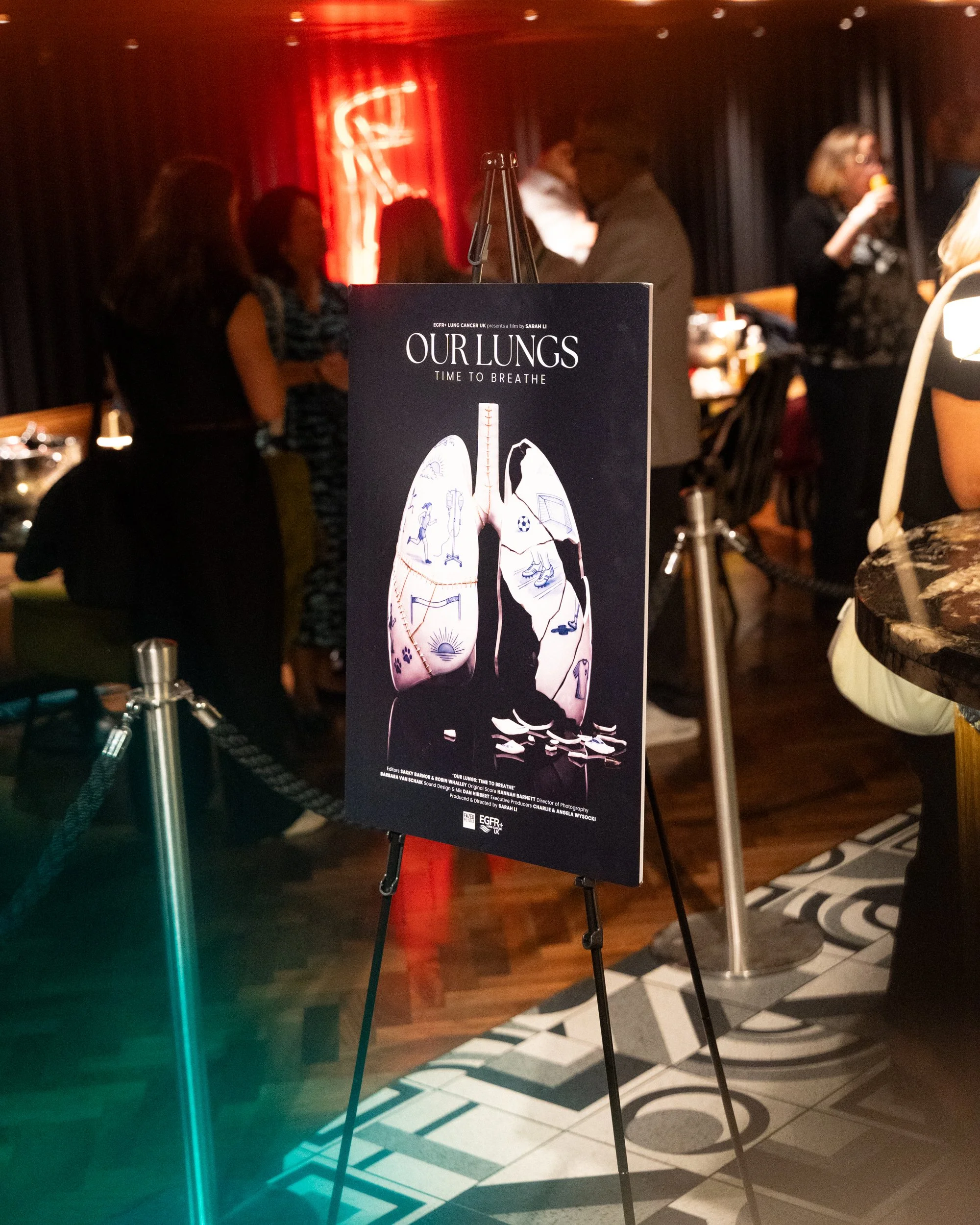 Poster for the film 'Our Lungs: Time to Breathe' in a dimly lit venue with people in the background, some standing and some seated, near a black and white patterned floor. The poster features an illustration of human lungs with smaller images inside 