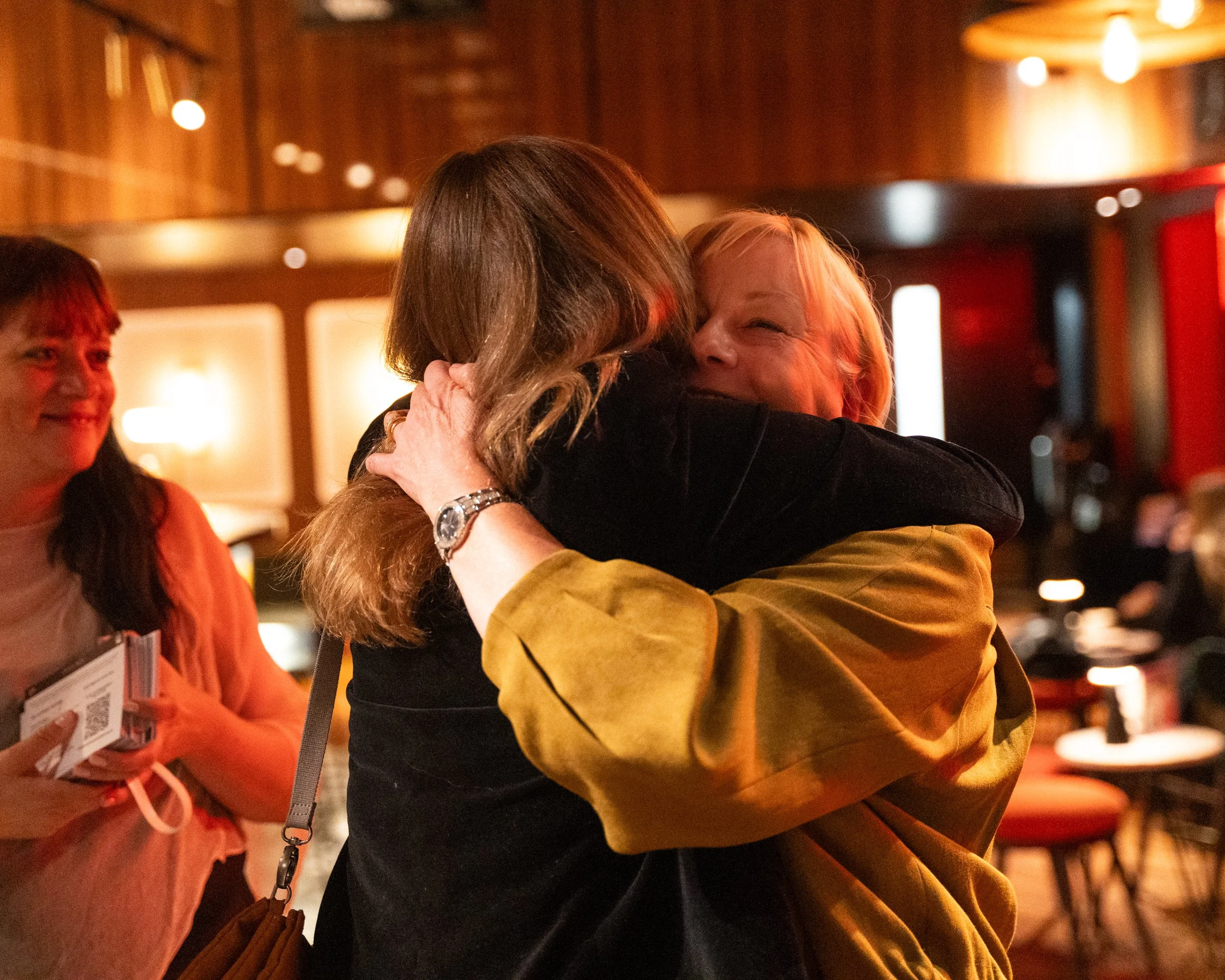 Two women are hugging each other warmly in a dimly lit indoor space, with one woman smiling happily and another with her face partially hidden. A third woman is standing nearby, holding a brochure or ticket, and smiling. The environment appears to be