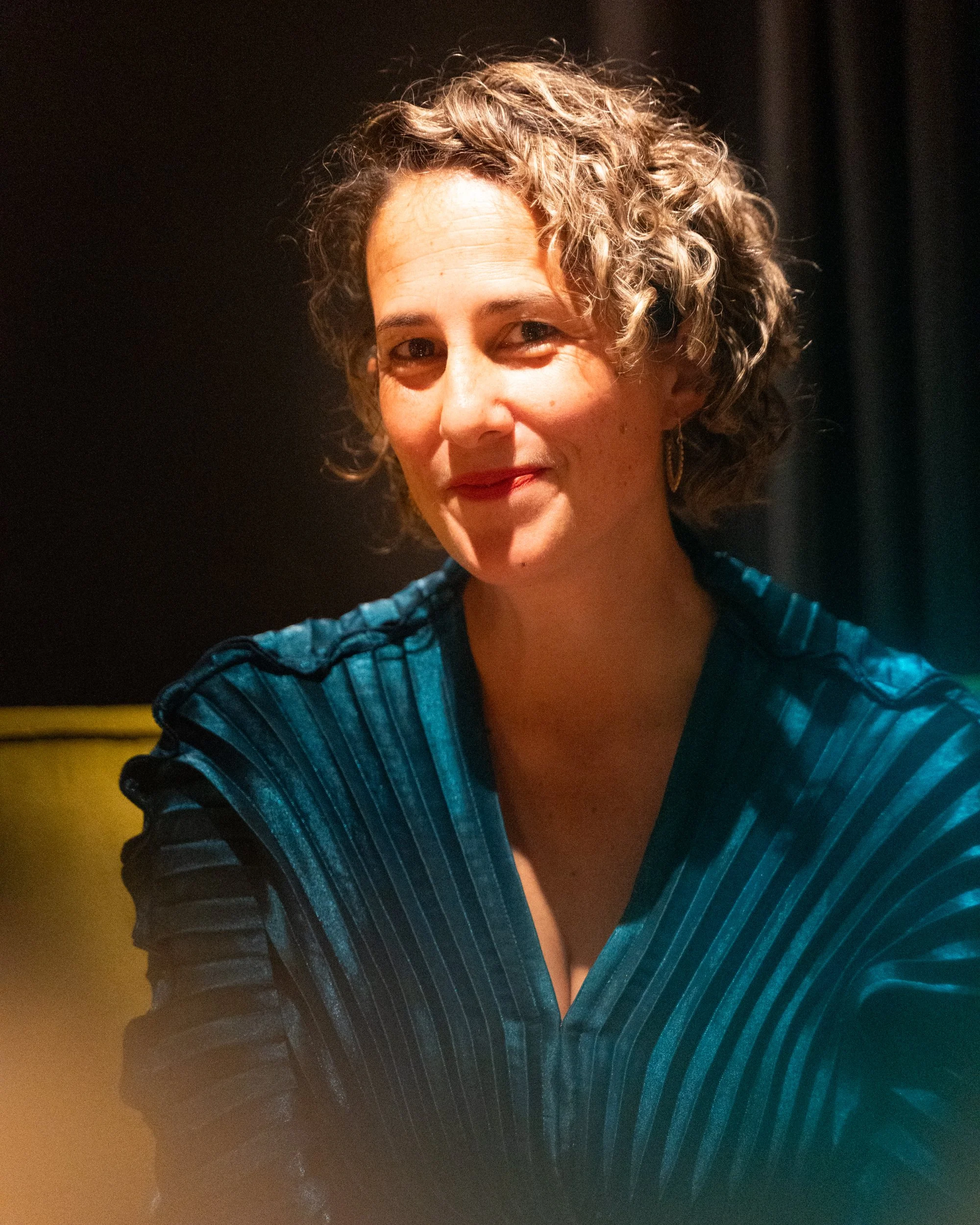 A woman with curly hair smiling in a dark setting, wearing a blue top with a V-neck.