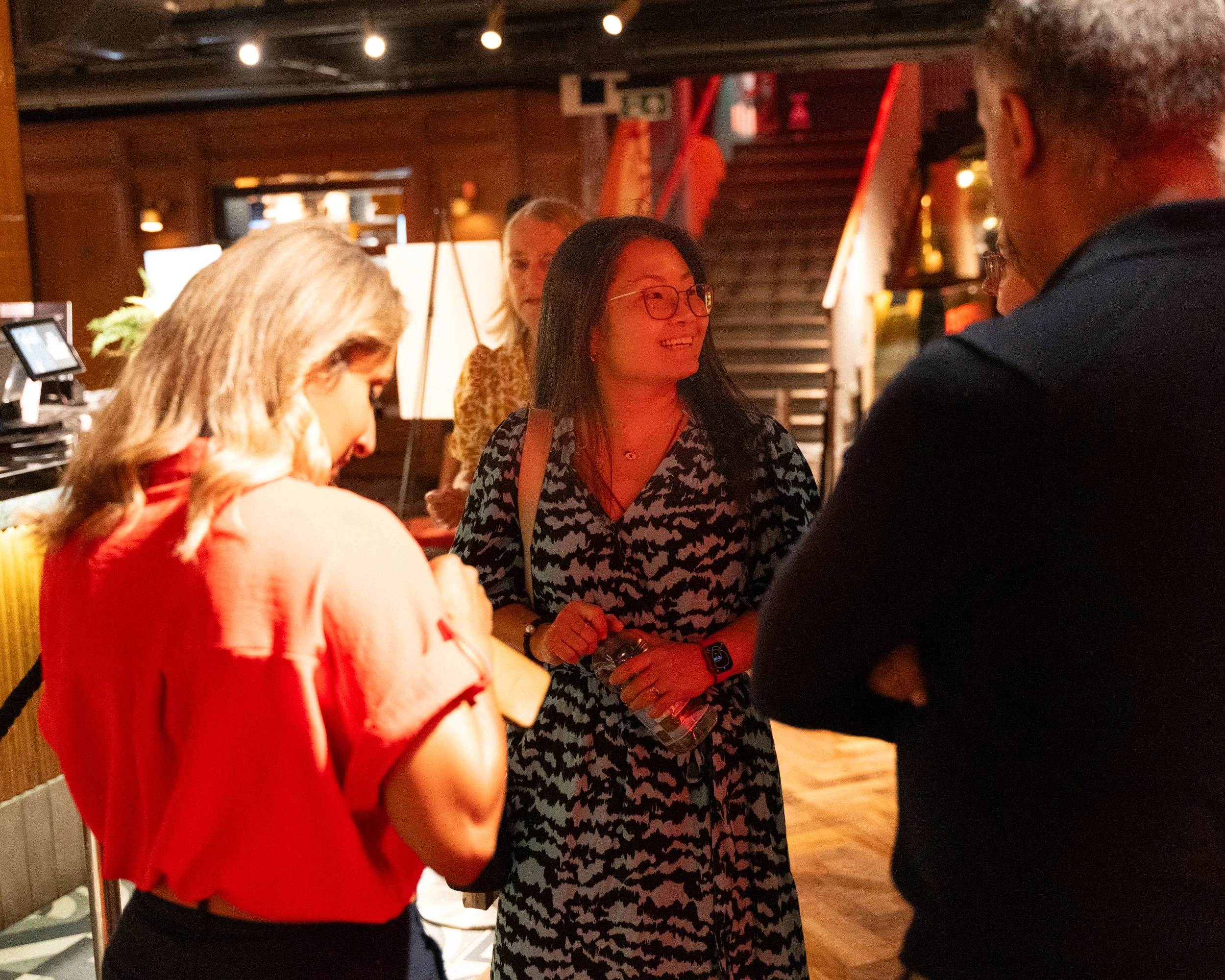 Group of people engaged in conversation in a warmly lit indoor setting, with dark wooden walls and staircase in the background.