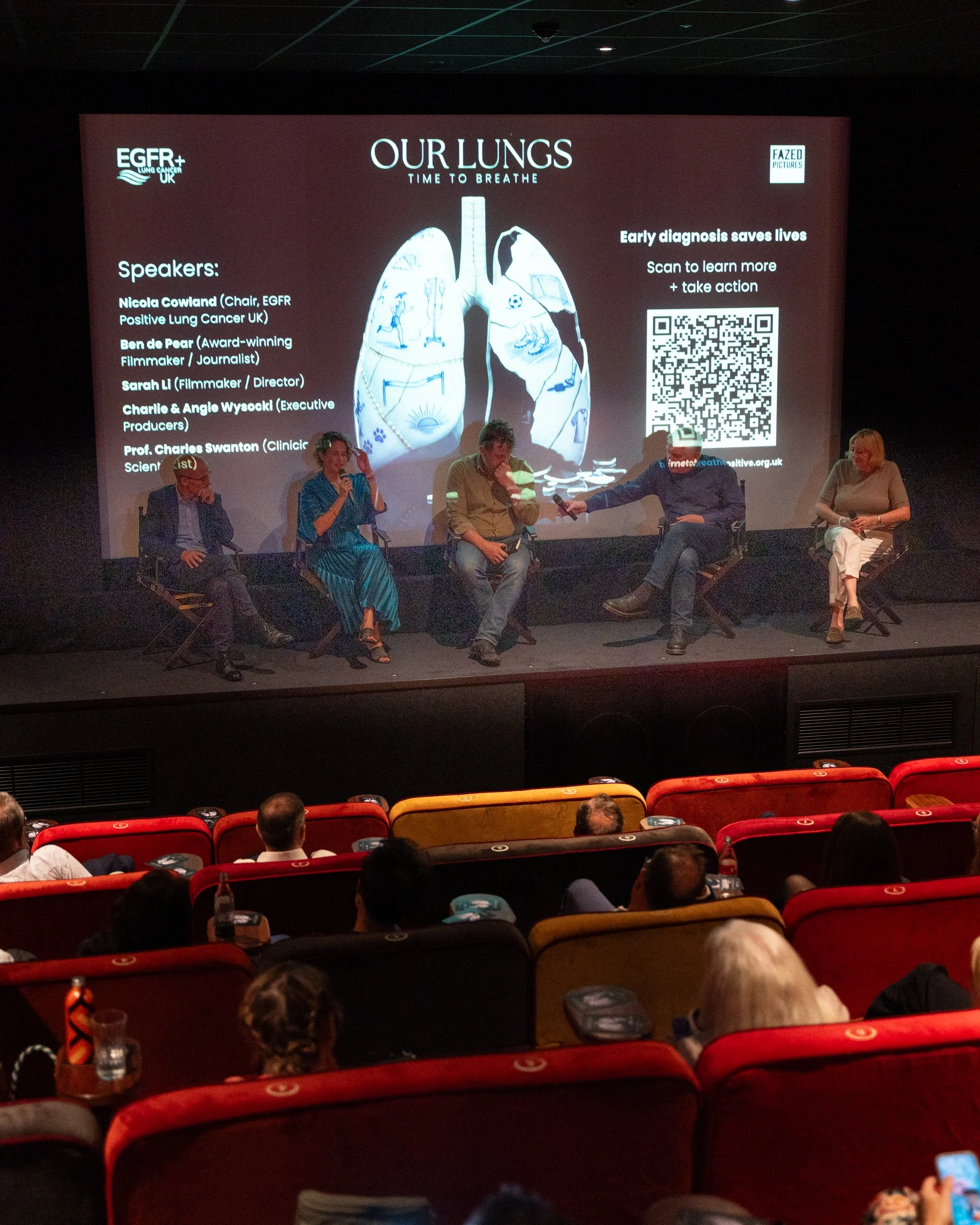 Panel discussion at a conference about lung health, with five panelists seated on stage, a large screen behind them displaying text and an image of lungs, and audience members in front.
