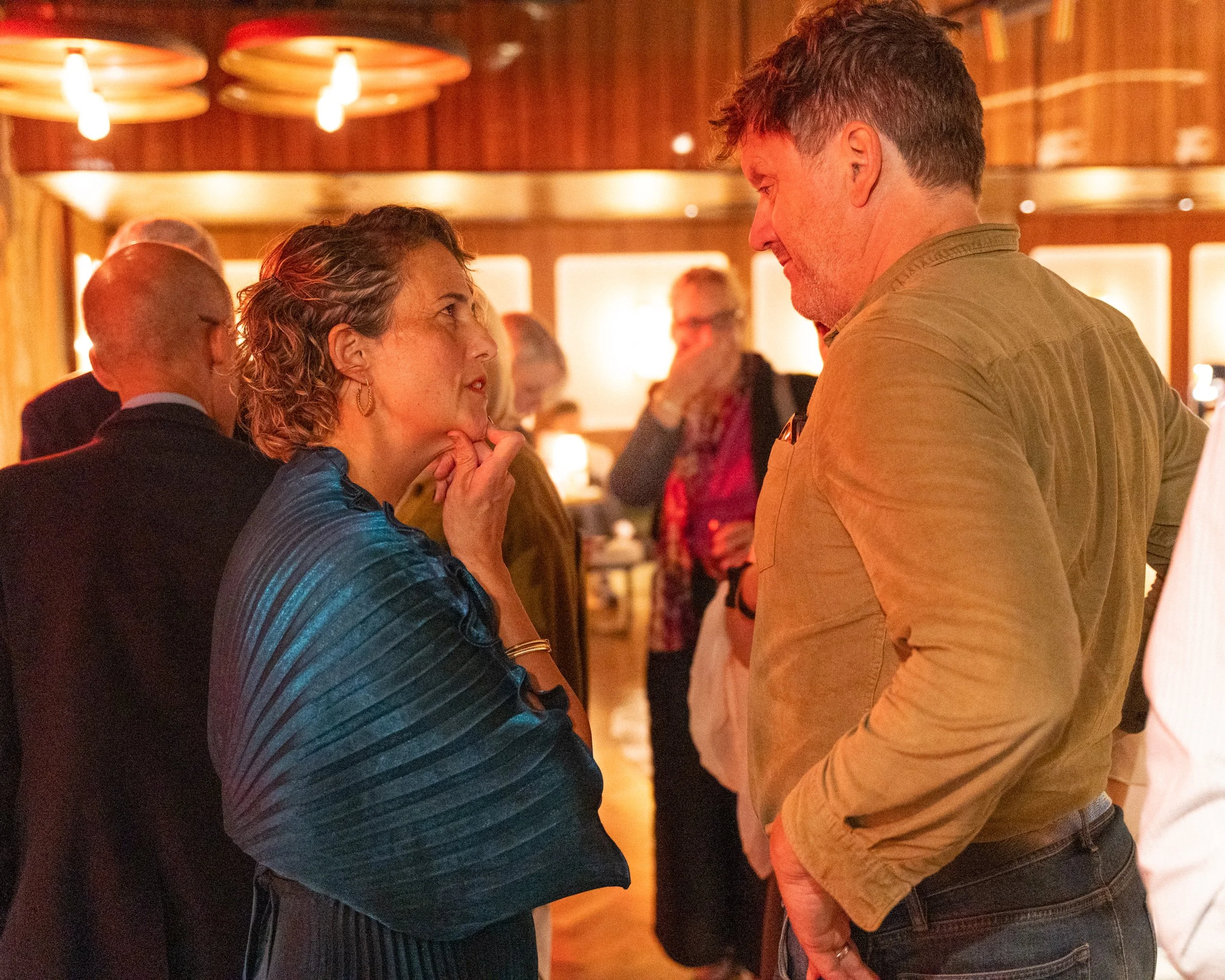 Two people engaging in a conversation at an indoor social event, with warm lighting and other guests in the background.