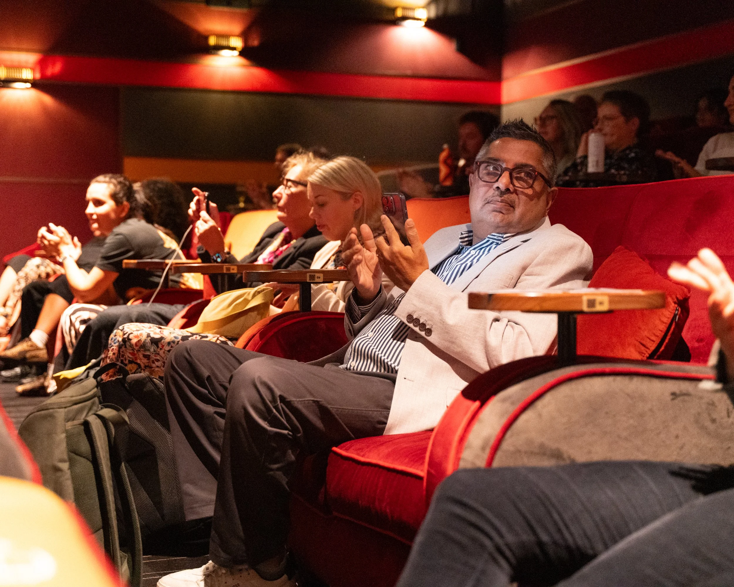 People seated in a theater or auditorium, some using their phones, with a man in the foreground wearing glasses and a light blazer.