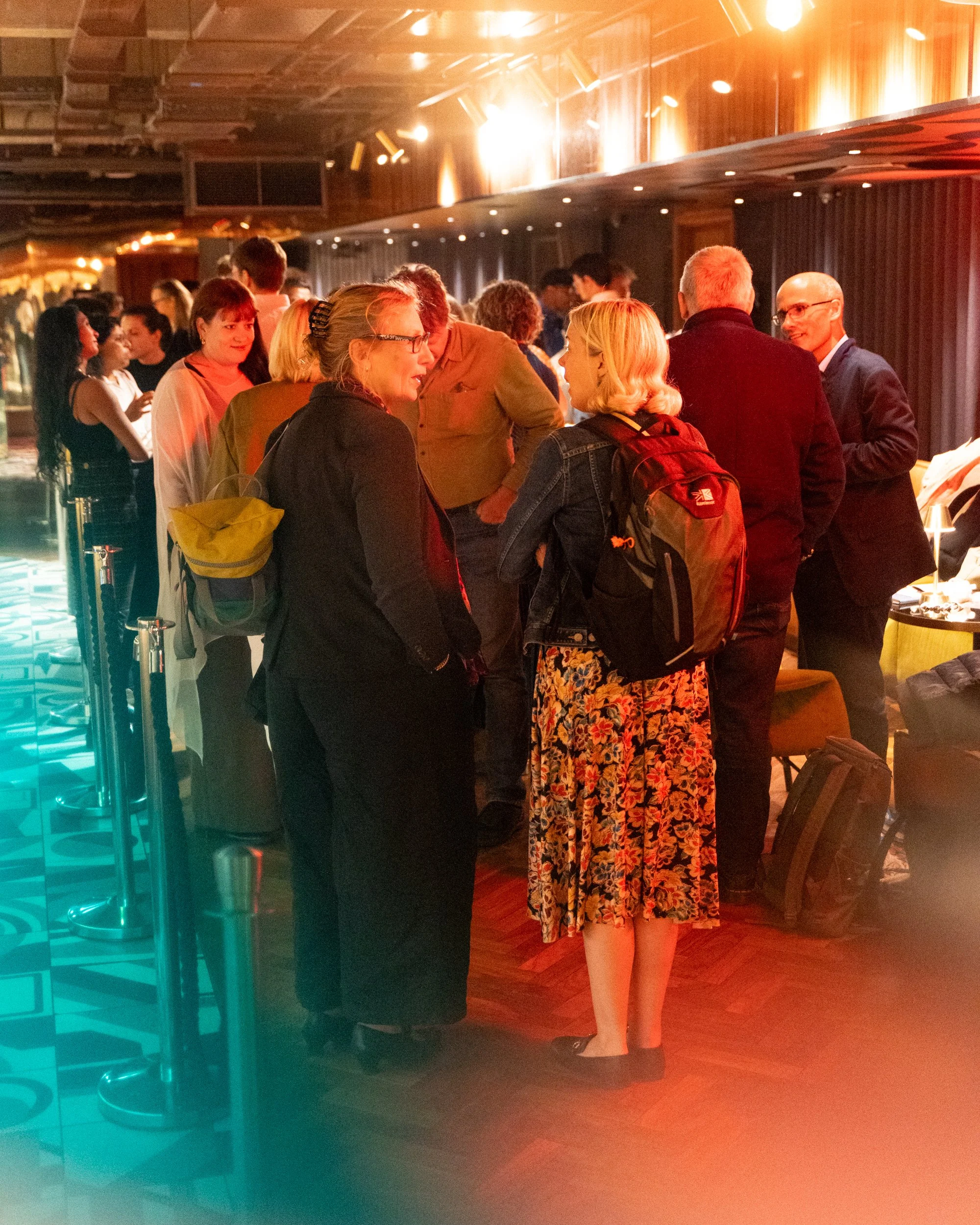 People gathered at an indoor event, engaged in conversations, with a woman in floral dress and others in business casual attire, dim lighting, and wooden interior.