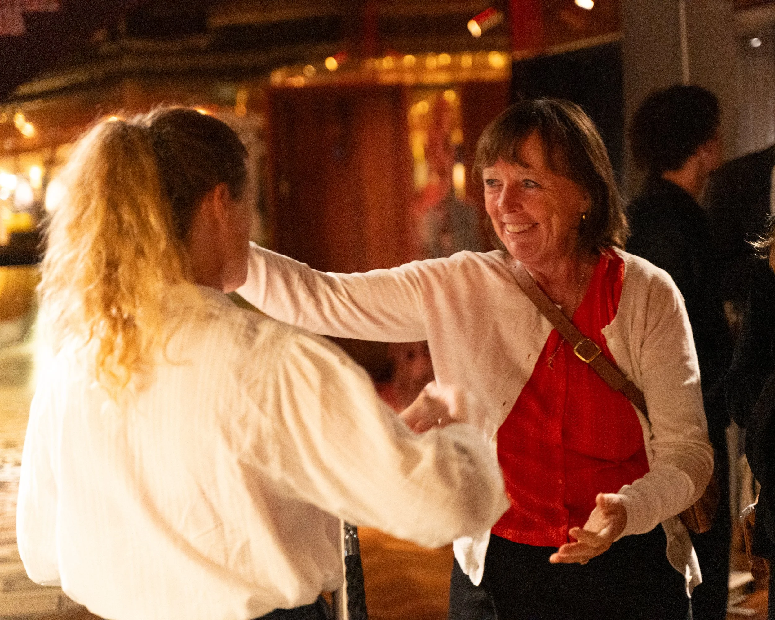 Two women smiling and interacting at a social gathering or party, with dim lighting and a blurred background.