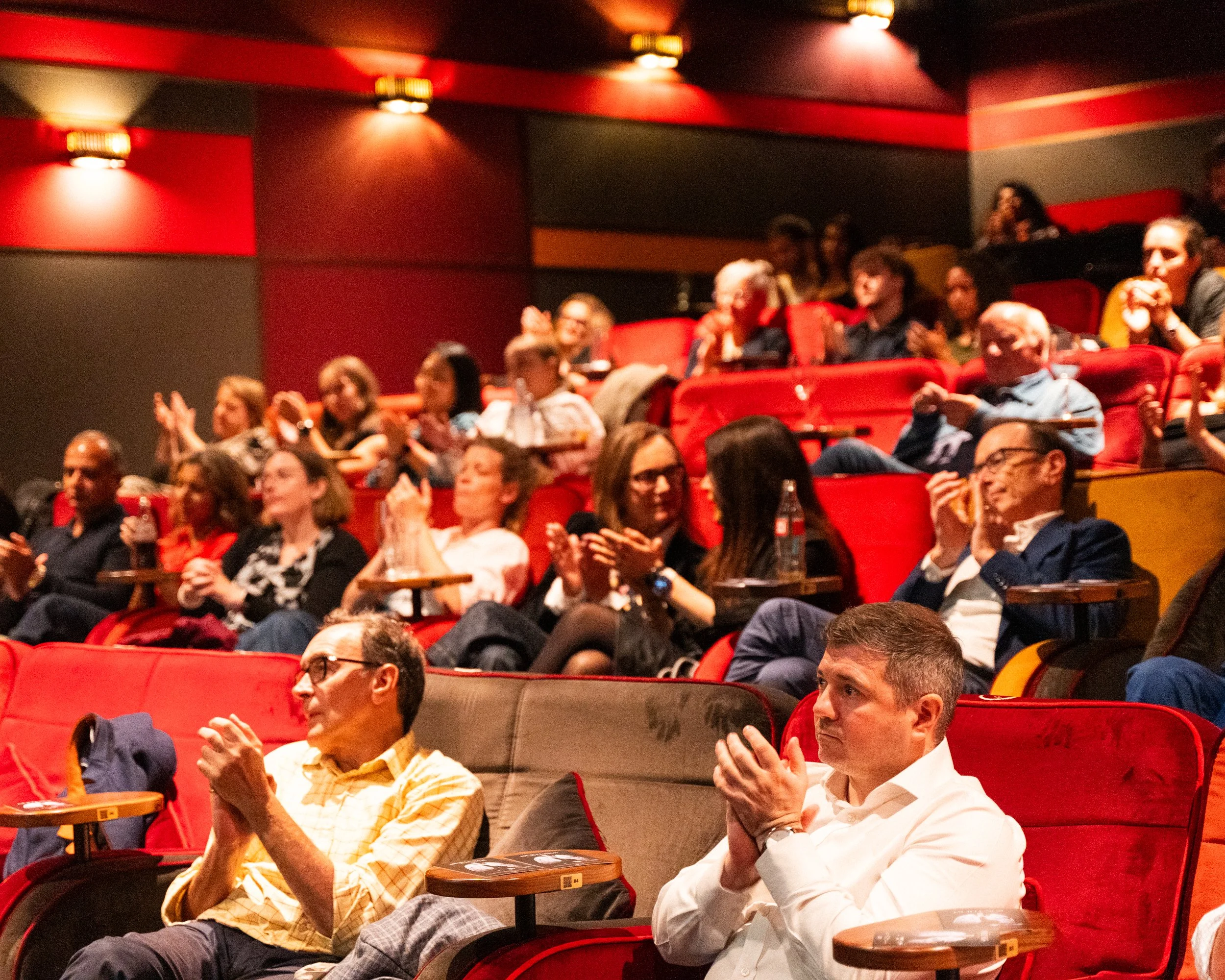 Audience seated in a theatre with red and black walls, some clapping and some taking photos.