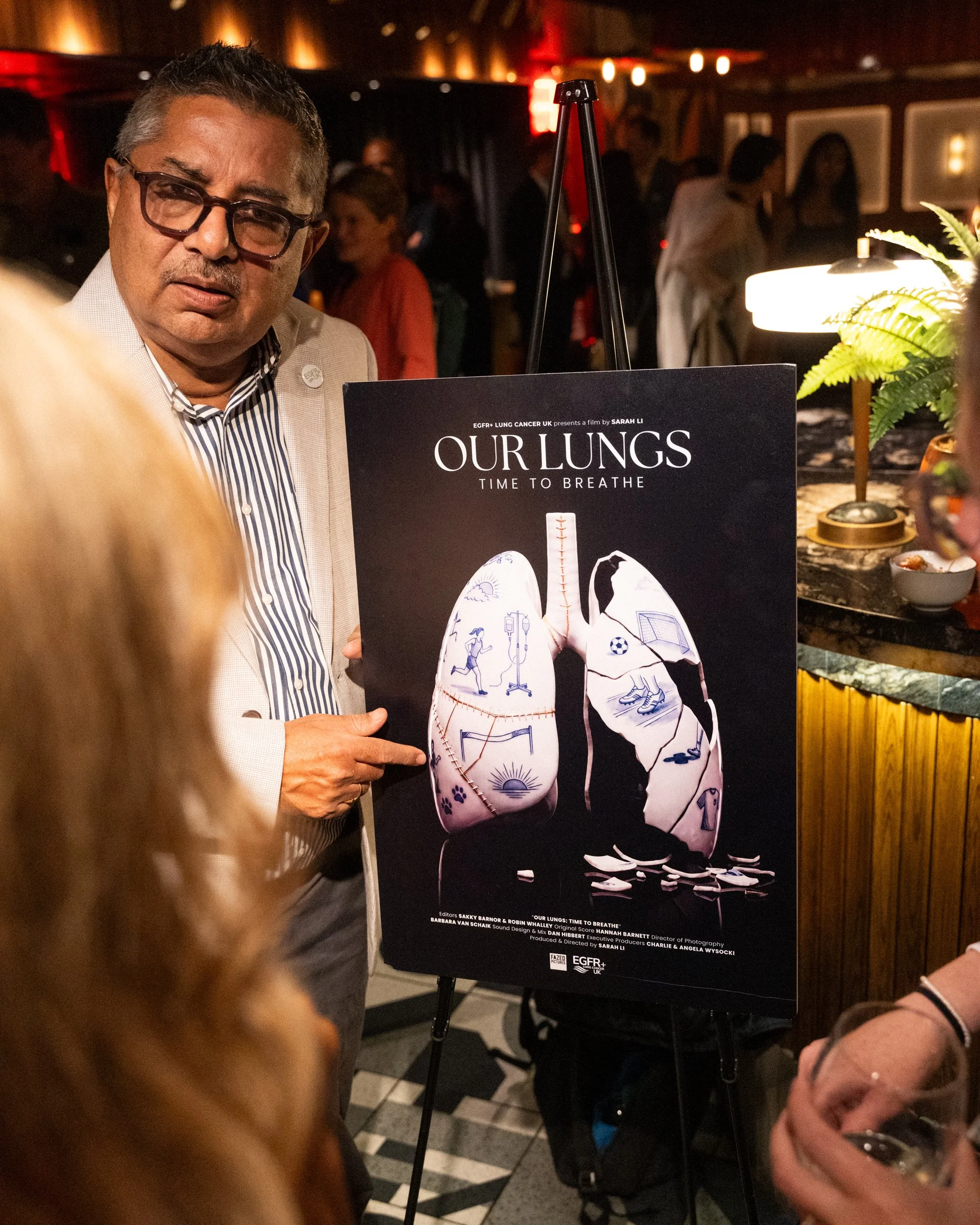 A man with glasses wearing a beige blazer and striped shirt standing next to a poster about lung health at an indoor event, with multiple people in the background.