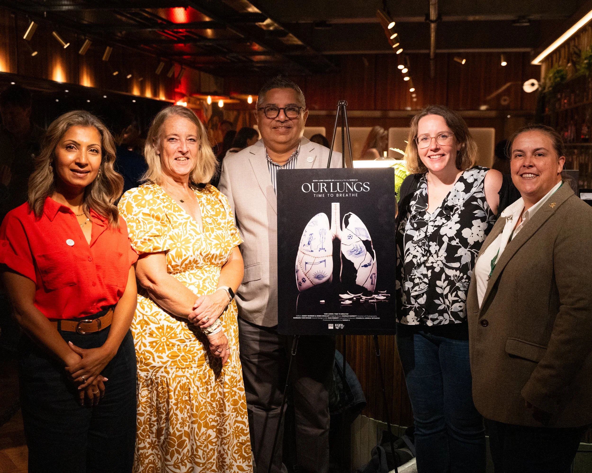 A group of six people posing in an indoor setting with dark wood walls and ambient lighting. There is a sign in front of them that reads 'Our Lungs: Time to Breathe,' featuring a diagram of lungs.