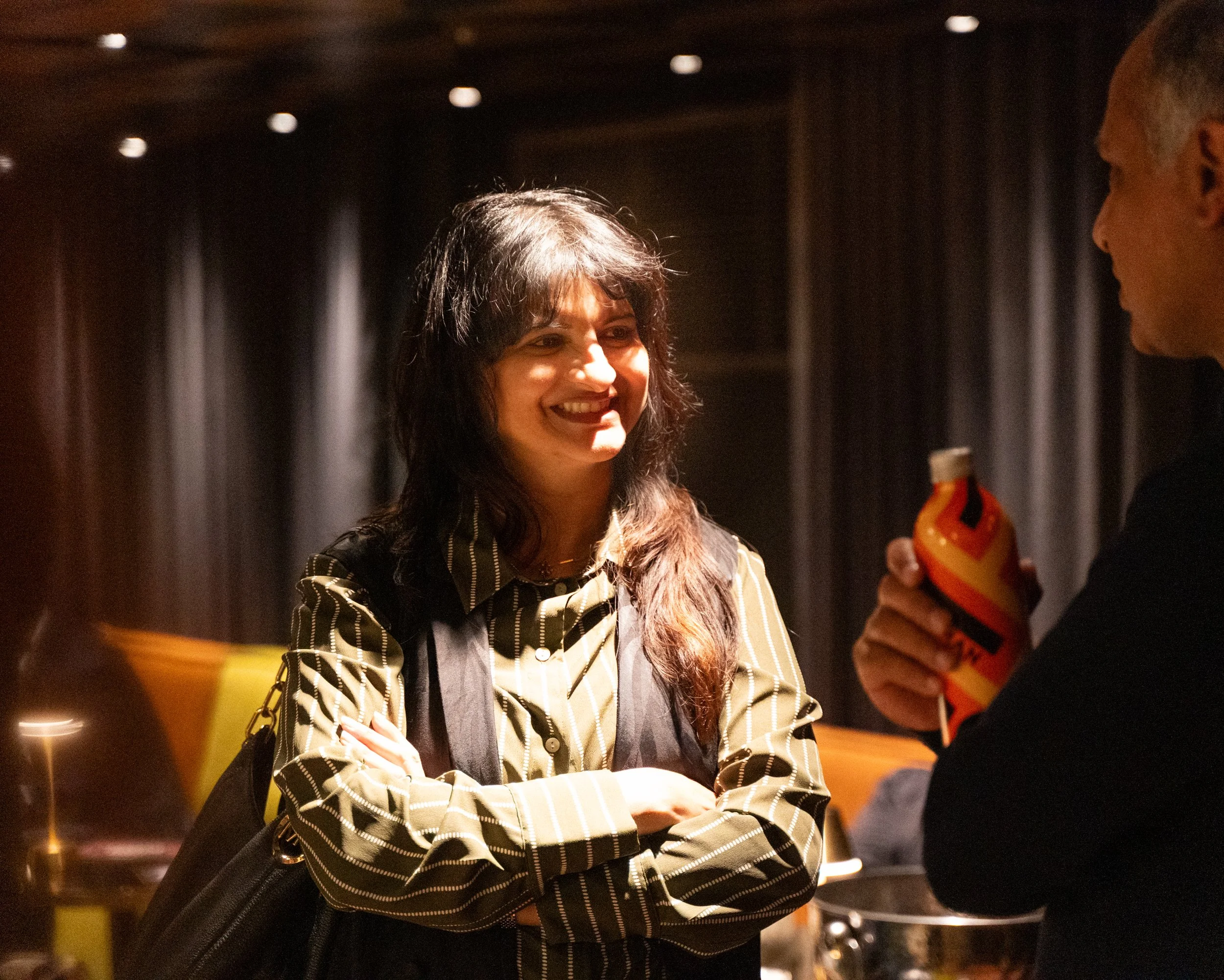 Two women are smiling and engaging in conversation inside a dimly lit room, with one woman wearing a black and white striped shirt and a black bag, and the other holding a colorful (red, yellow, black) object.