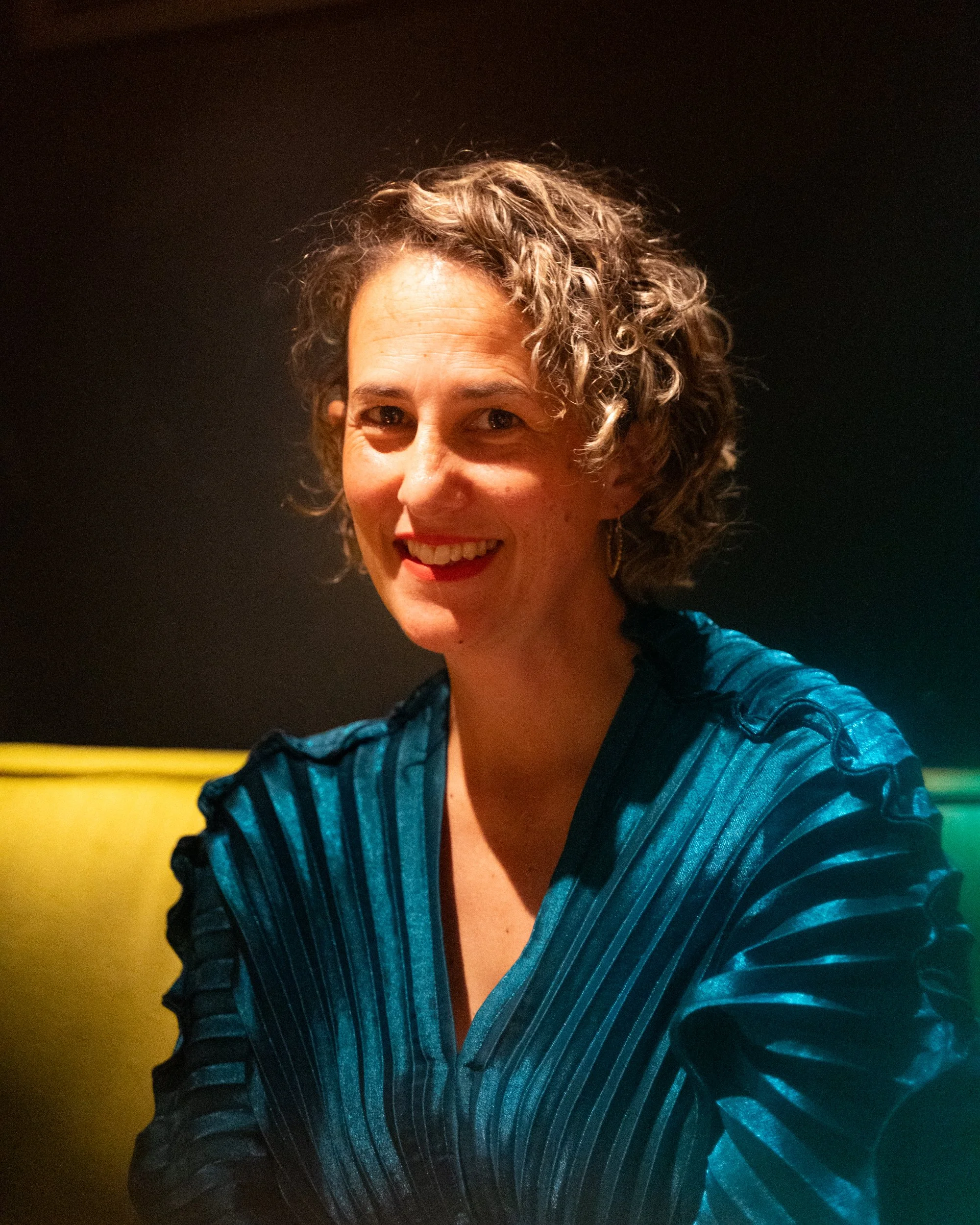 A woman with short curly hair taking a selfie in a dimly lit room. She has a smile and is wearing a shiny blue jacket with black accents.