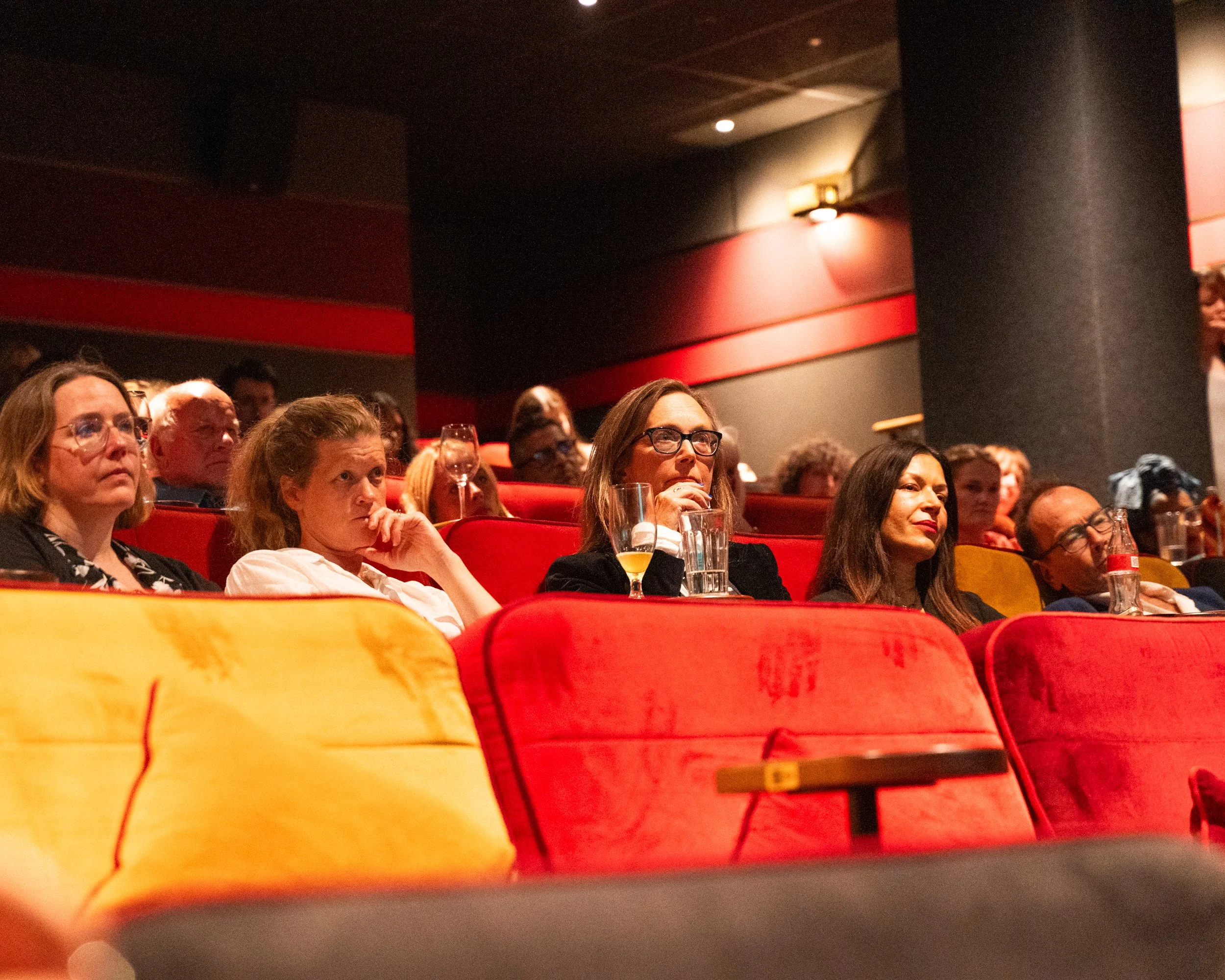 Audience seated in a theater watching a presentation or performance, with red and yellow theater seats and dim lighting.