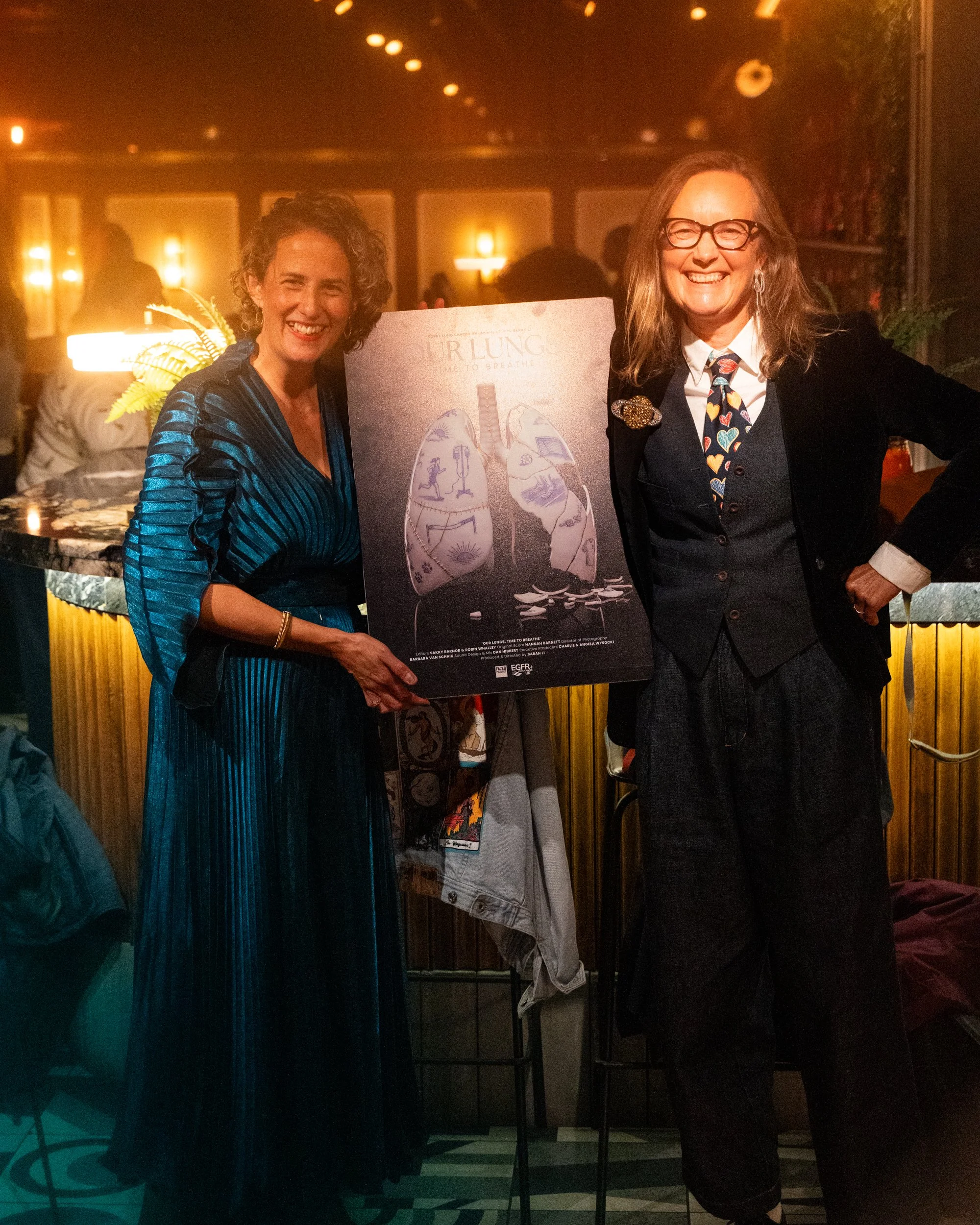 Two women smiling and posing for a photo in a dimly lit room, with one holding a poster depicting lungs with illustrations inside them. The woman on the left wears a shiny blue dress, and the woman on the right wears glasses and a black blazer with a