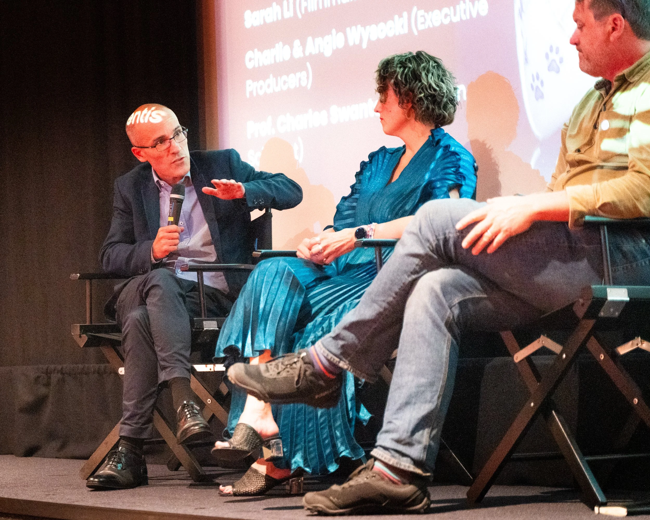 A panel discussion at an event with three people on stage, two men and one woman, sitting on stage chairs. One man is speaking into a microphone, the woman is listening attentively, and the other man is looking at the speaker. A large screen behind t
