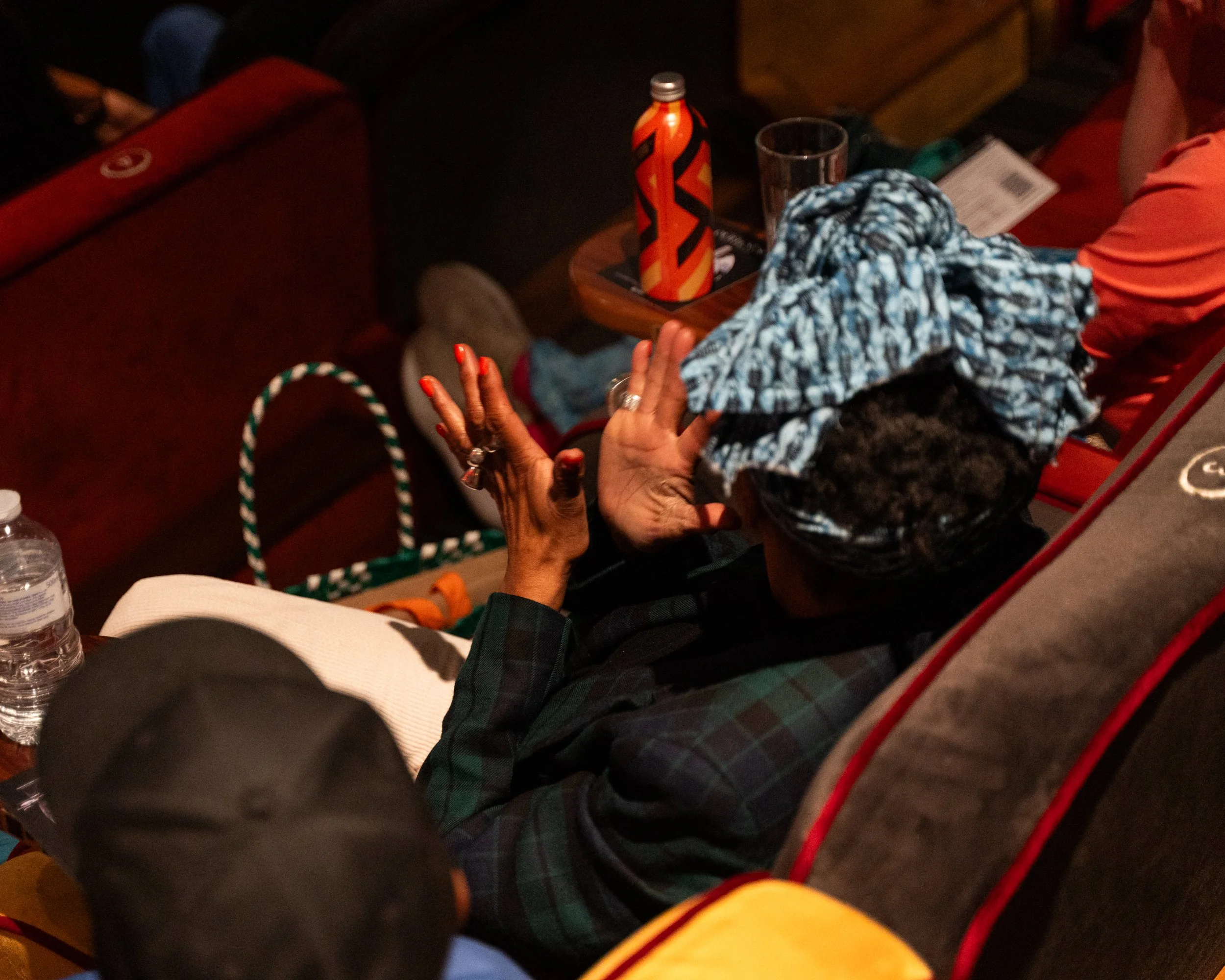 Close-up of a person with dark curly hair and painted red nails, holding hands with a child who has dark skin and is wearing a blue and white patterned headscarf, in a dimly lit setting with a table in the background.
