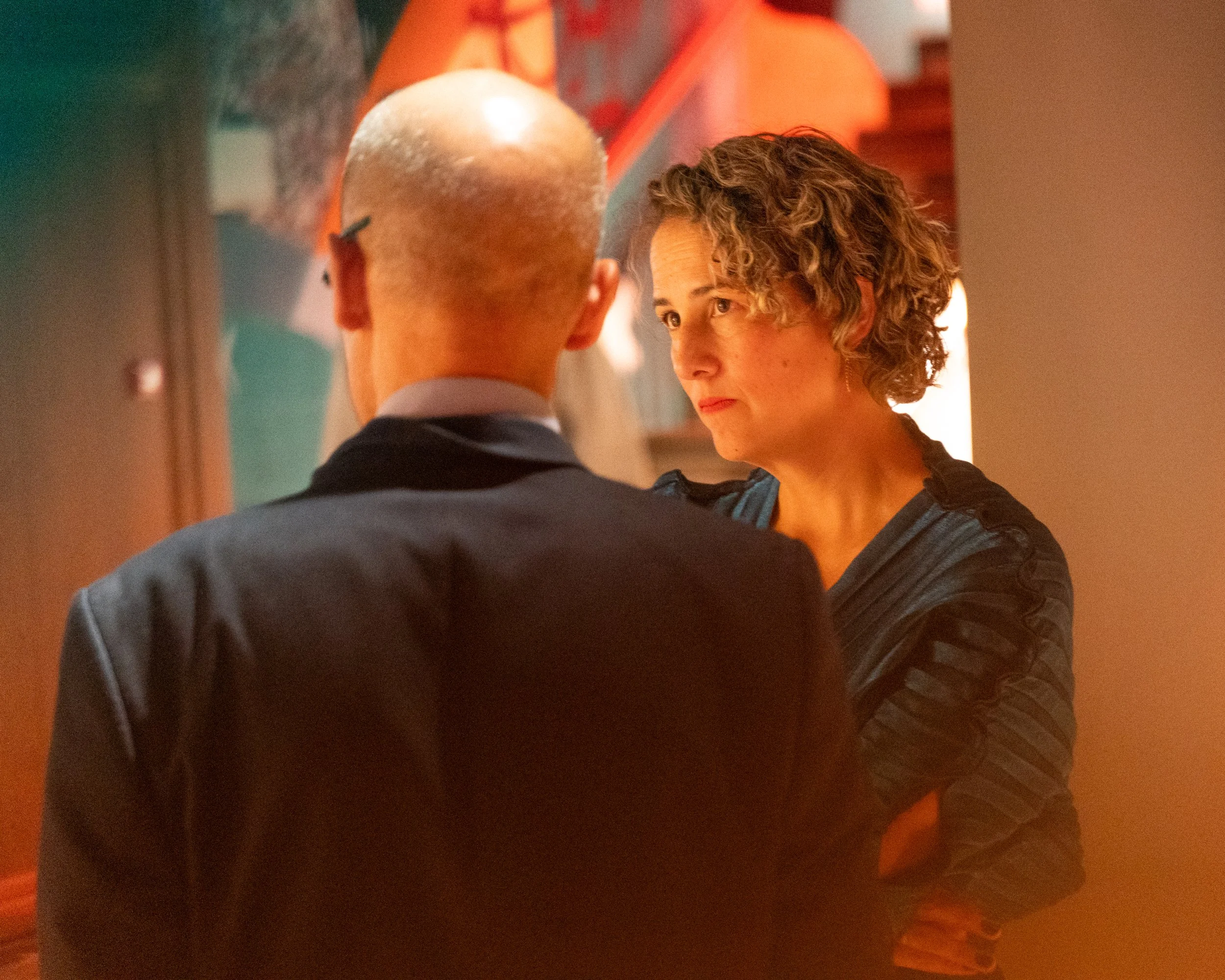 A man and woman face each other closely indoors, with the woman looking into the man's eyes. The background has warm lighting and art decor.