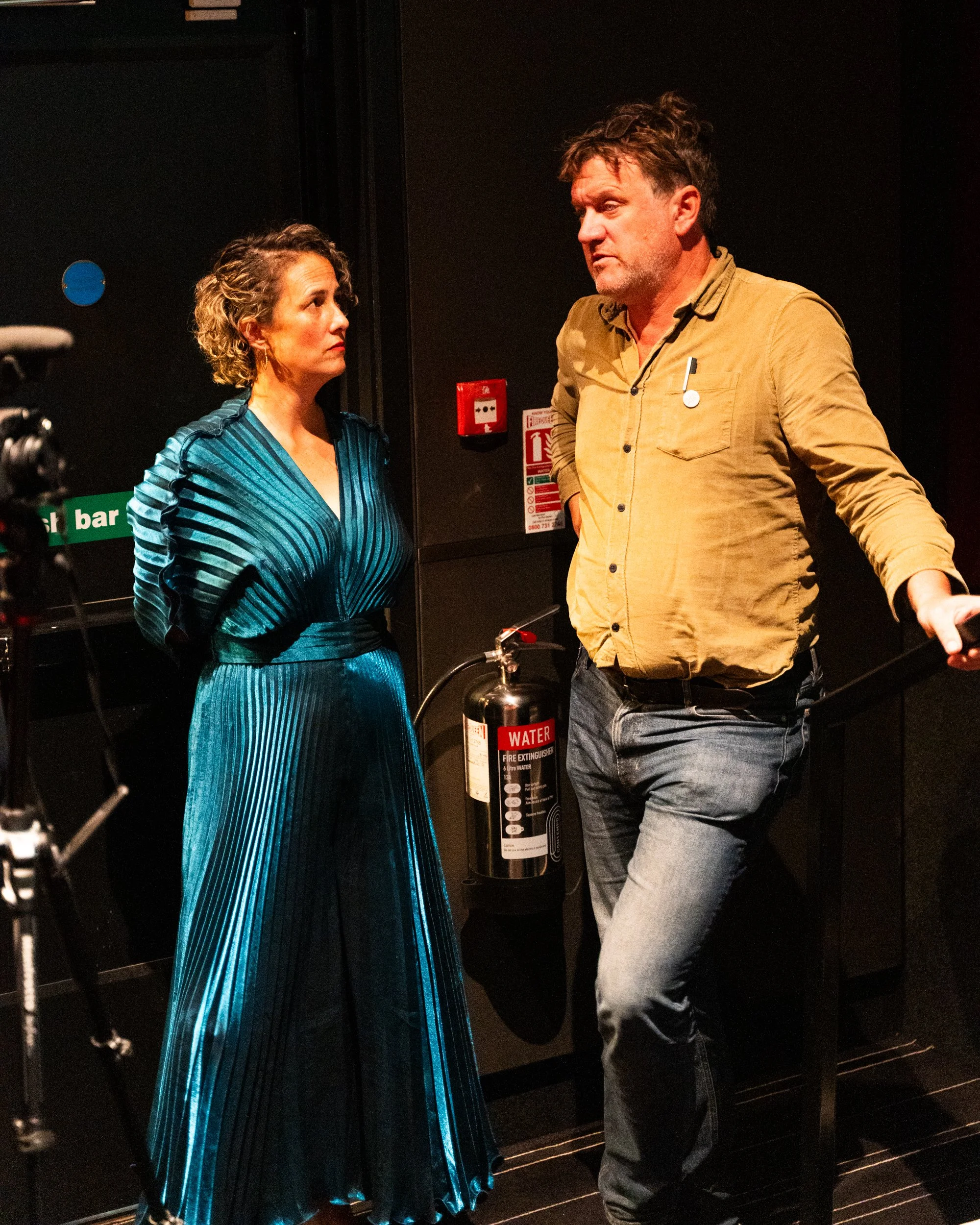 A woman and a man engaged in conversation in a dimly lit room. The woman is wearing a shiny blue dress and has curly hair, while the man is dressed in a beige shirt and jeans. There are safety signs and a fire extinguisher visible in the background.
