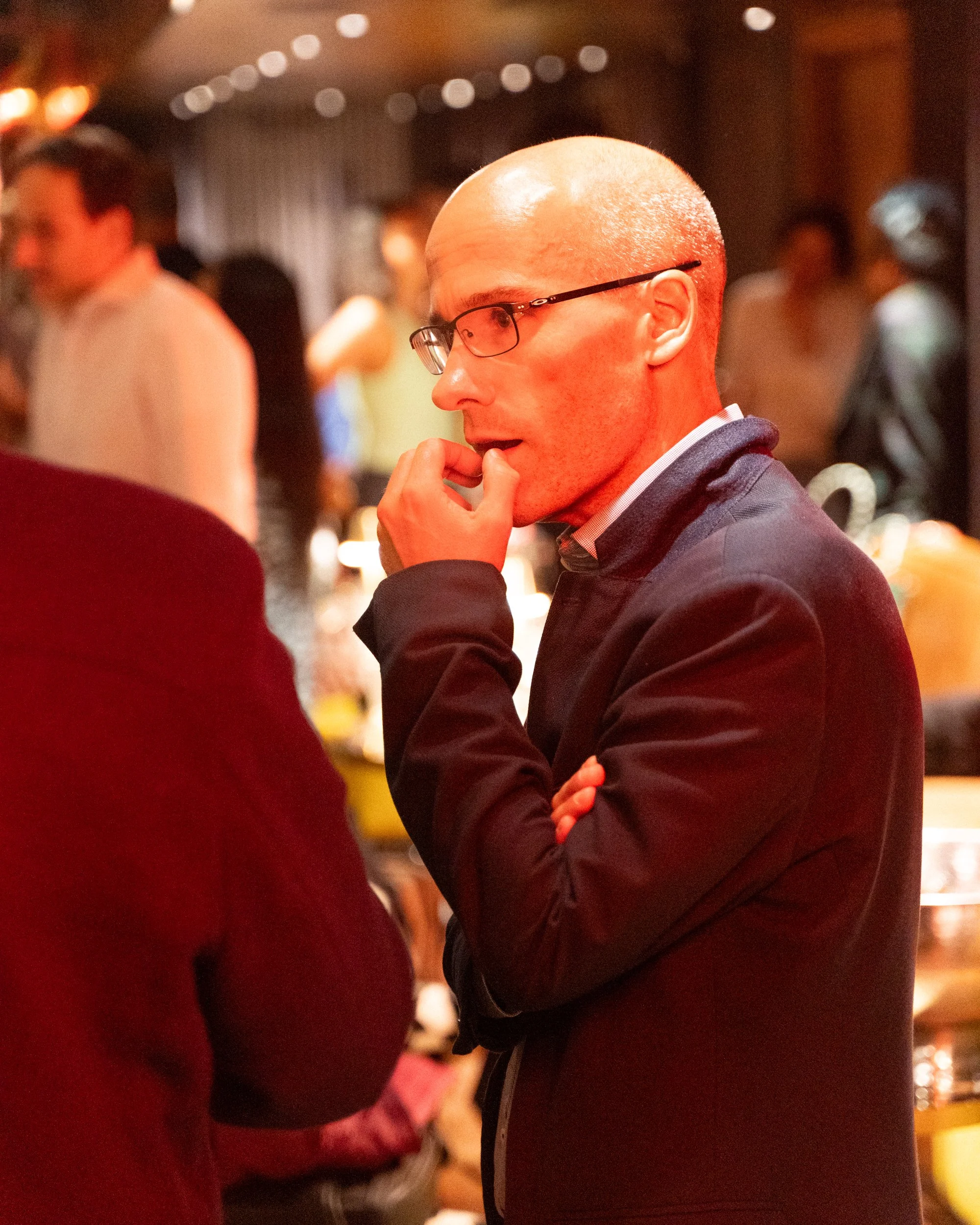 A close-up side profile of a bald man wearing glasses, a dark blazer, and a shirt, with hand on chin, in a dimly lit social setting with people in the background.
