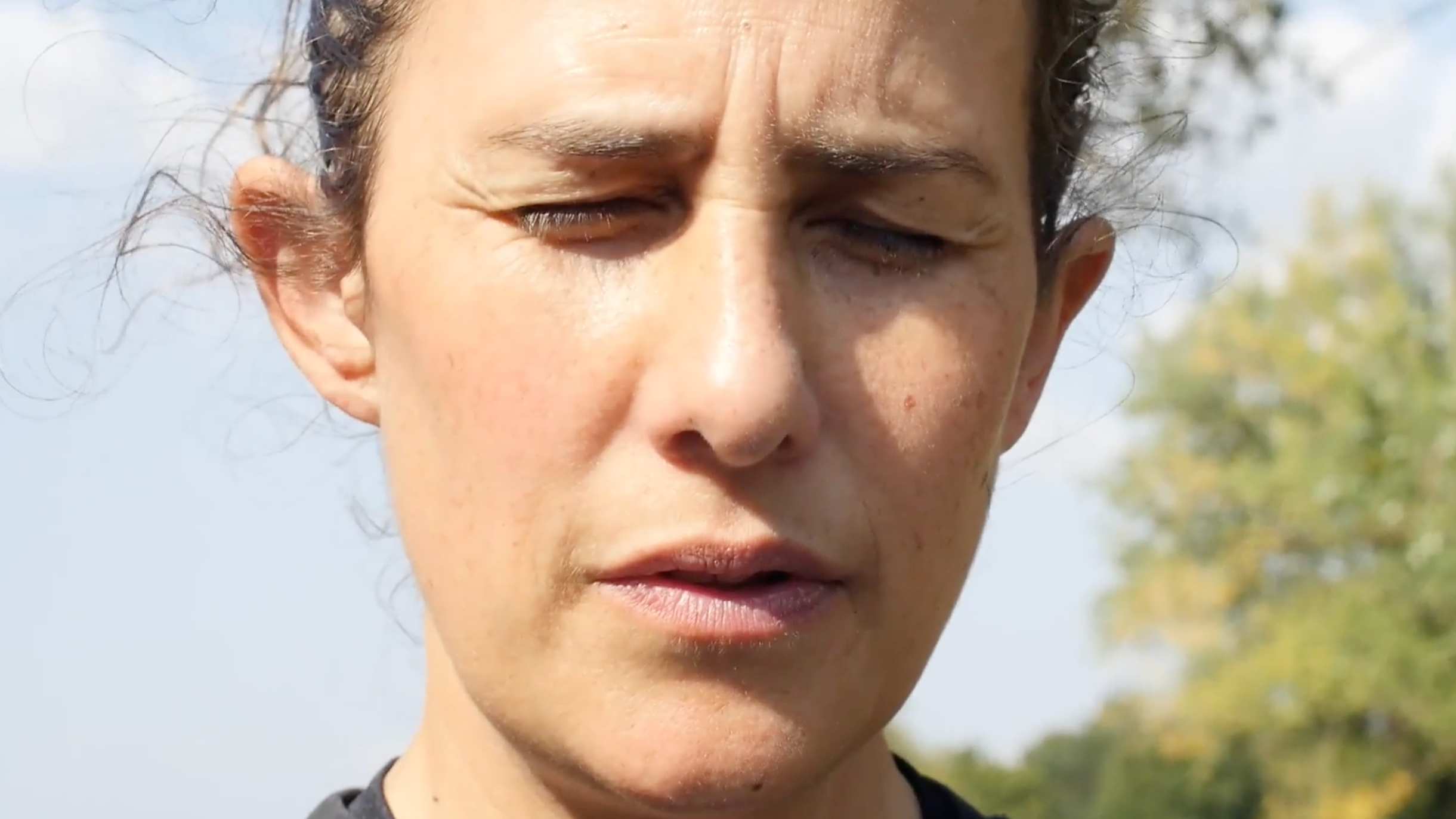 A close-up of a Sarah Li's, the filmaker of Our Lungs: Time to Breathe, face with eyes closed, outdoors on a sunny day, with blurred green trees in the background.