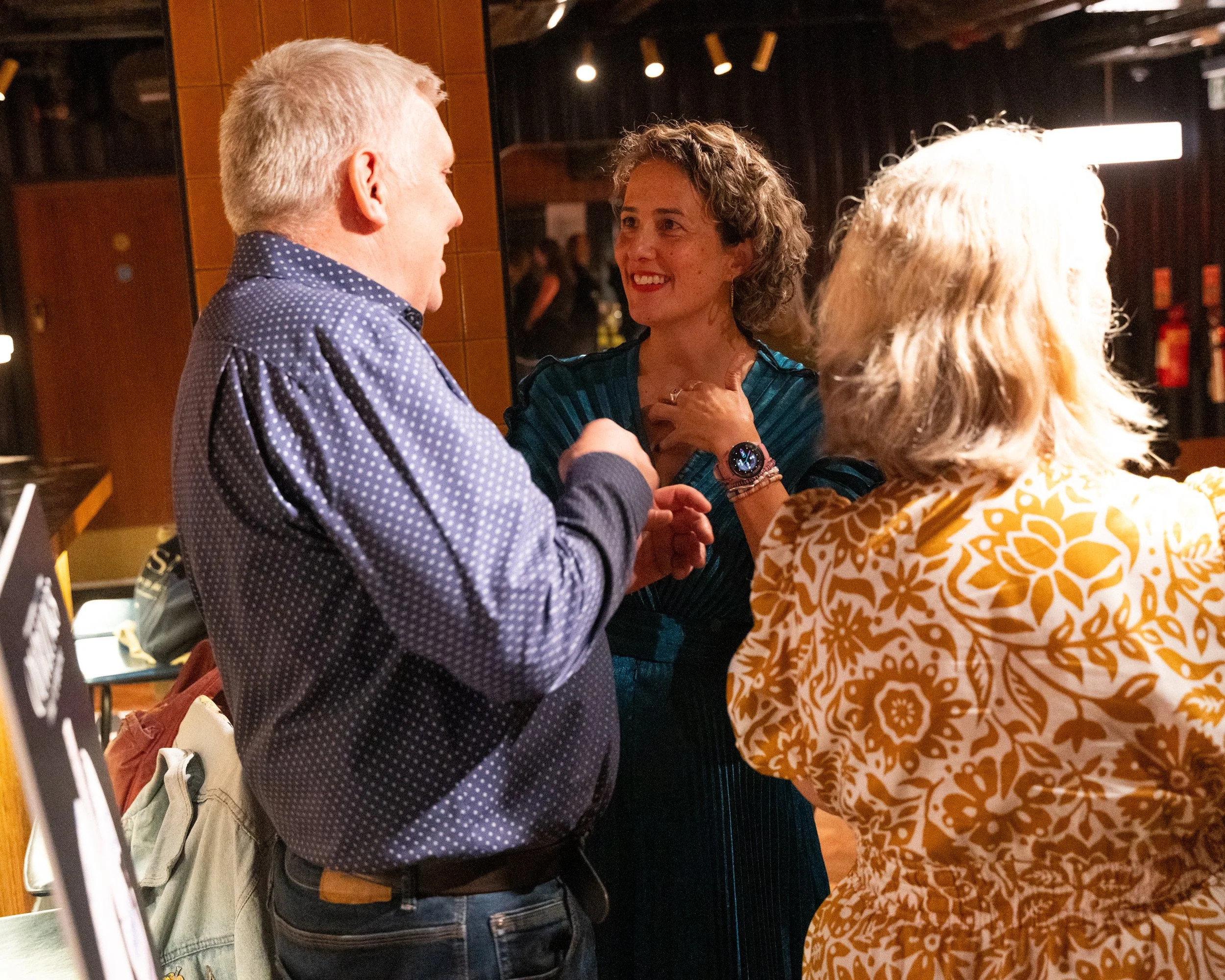 Three people engaged in conversation inside a warmly lit indoor space, with one woman smiling and touching her chest while talking to two men, one wearing a blue polka-dot shirt and carrying a backpack.