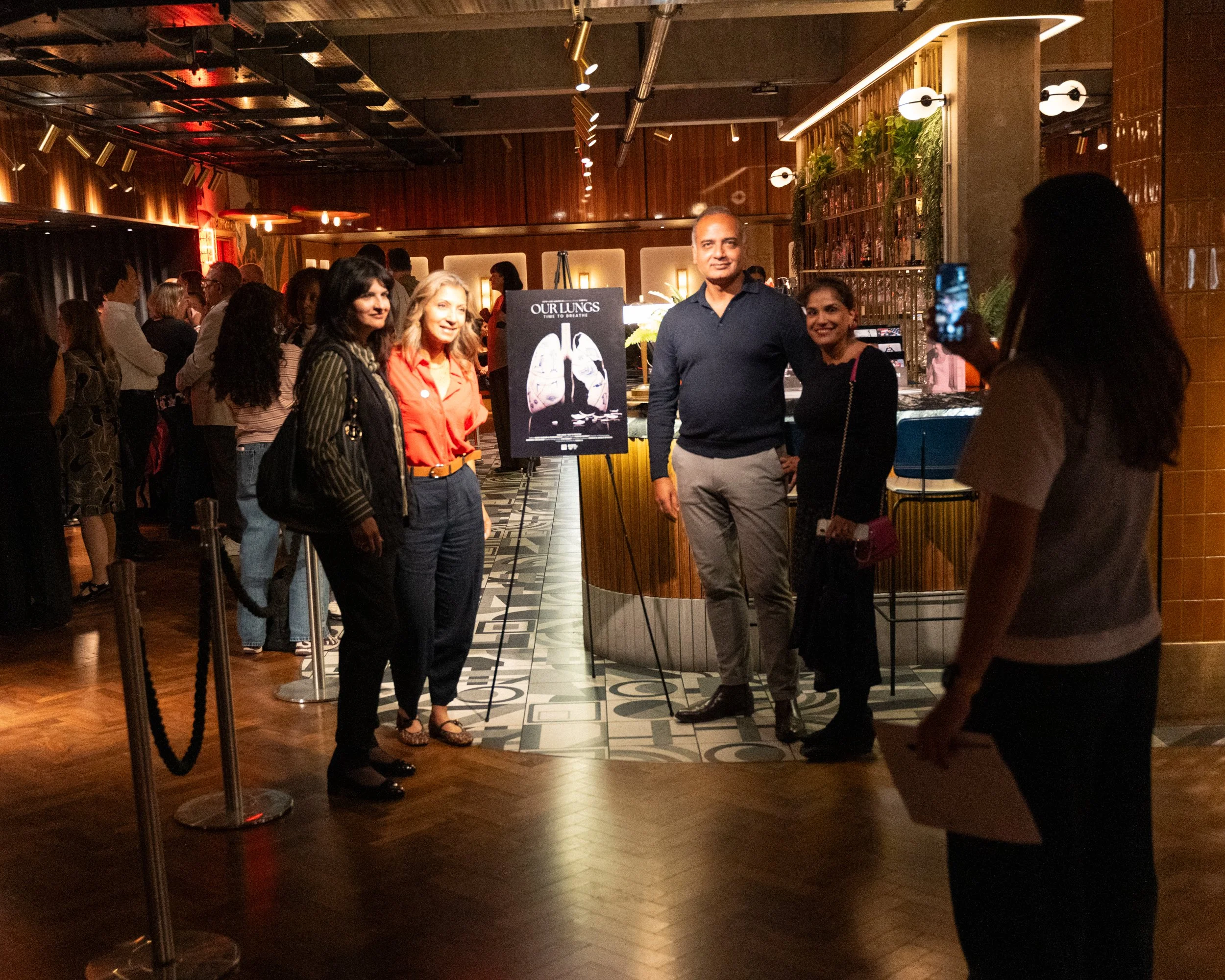 People standing in a dimly lit bar or restaurant, taking photos and socializing, with a sign that reads "Our Lungs: Time to Breathe."