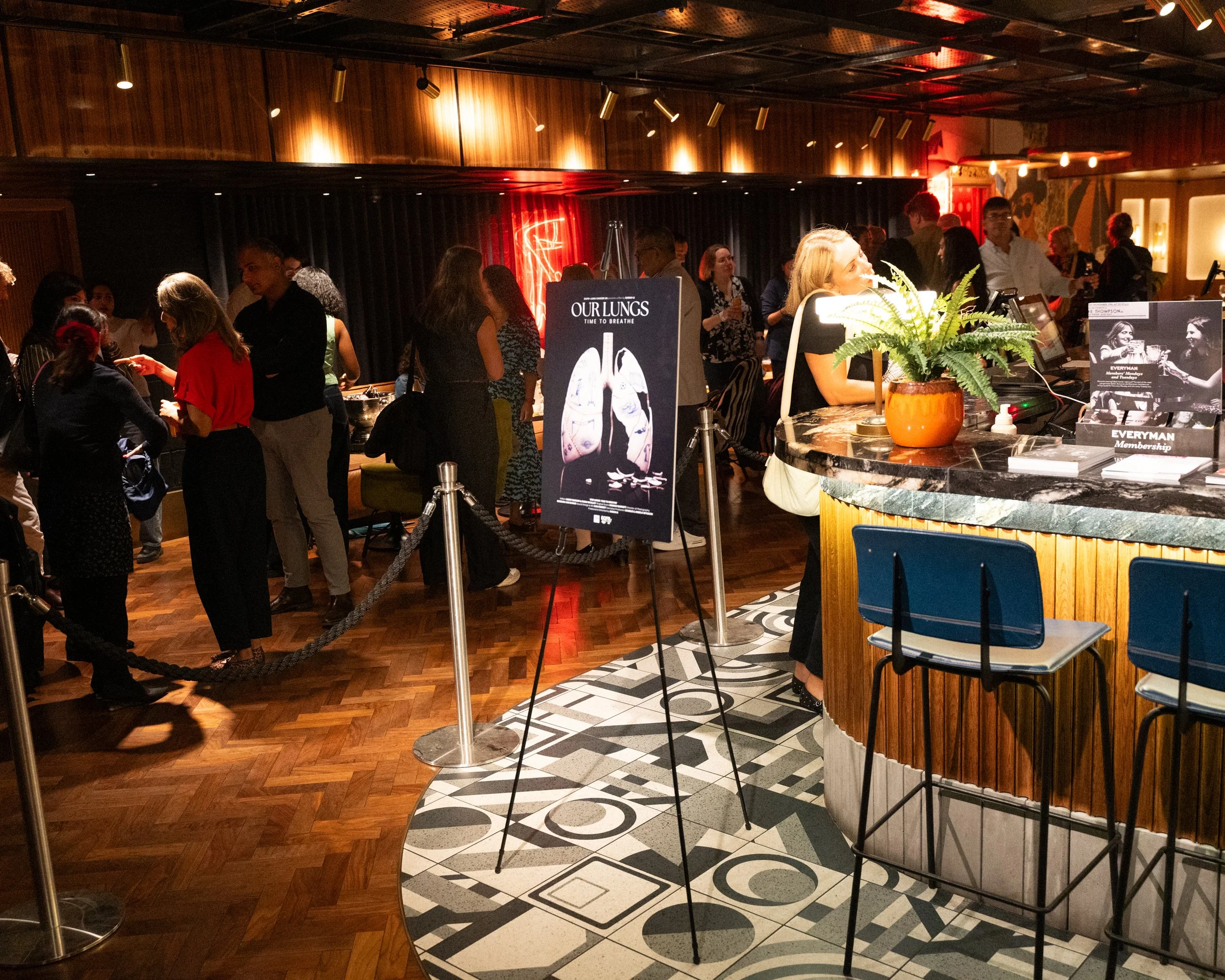 A crowded indoor event space with people standing and talking. There is a bar counter with a potted fern plant and promotional signs. The background features wood-paneled walls, warm lighting, and a dark curtain backdrop.