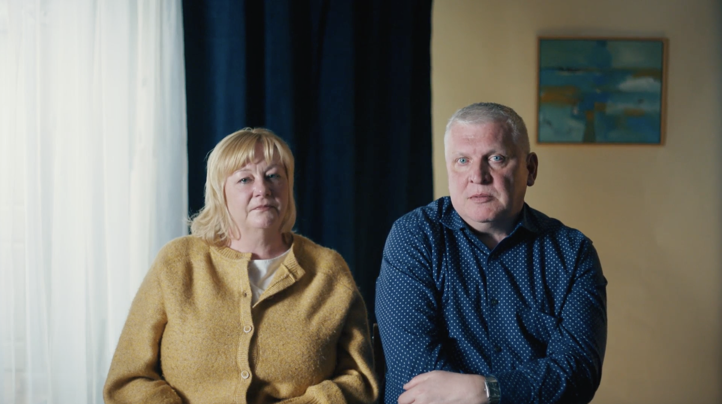 A middle-aged woman with blonde hair wearing a yellow cardigan and a middle-aged man with gray hair and blue eyes wearing a blue polka-dot shirt sit indoors near a window with light-colored curtains. A framed painting of a tree and sky hangs on the wall behind them.