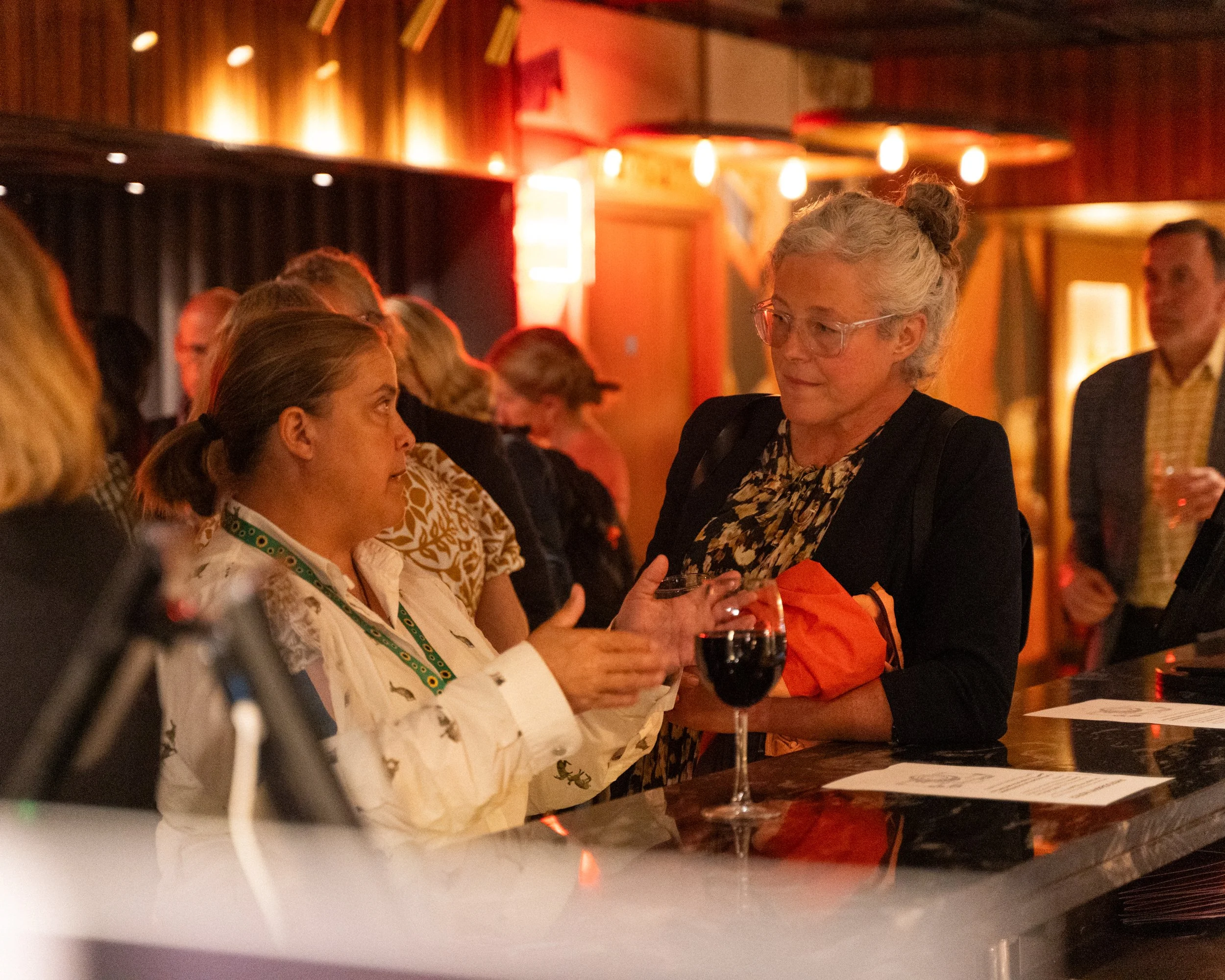 People socializing in a warmly lit bar or lounge, with wooden decor, some holding glasses of red wine, engaged in conversation.