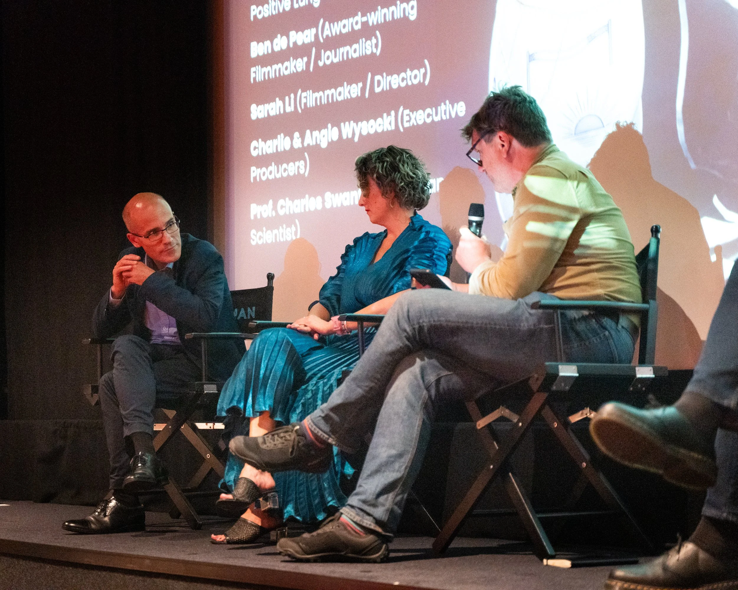 Three individuals seated at a panel discussion or interview on stage. A man on the left with glasses and a suit appears to be listening, a woman in the middle with short, curly gray hair and a teal outfit is looking downward, and a man on the right w