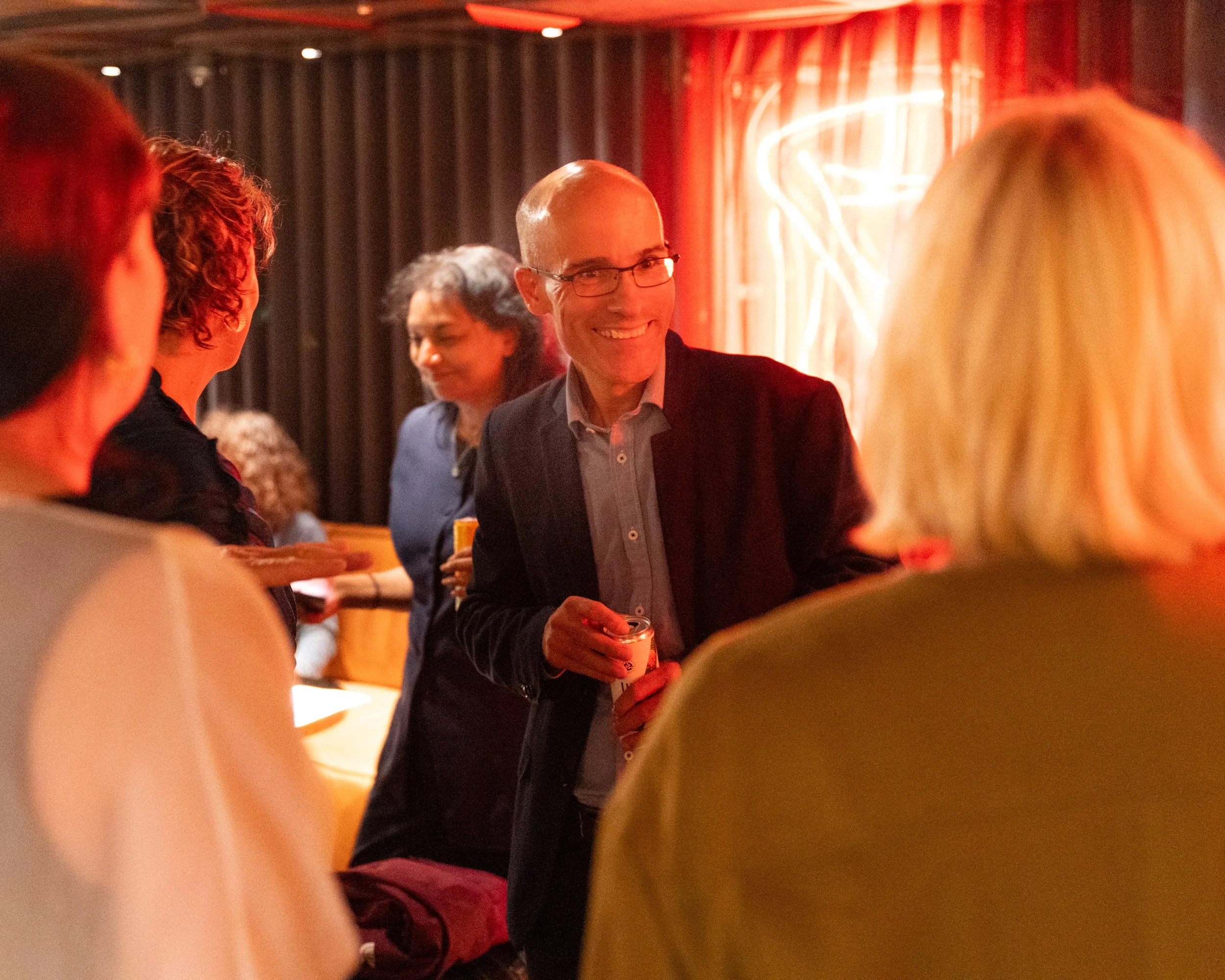 People socializing at a bar or restaurant, illuminated by warm red lighting, with a neon sign in the background.