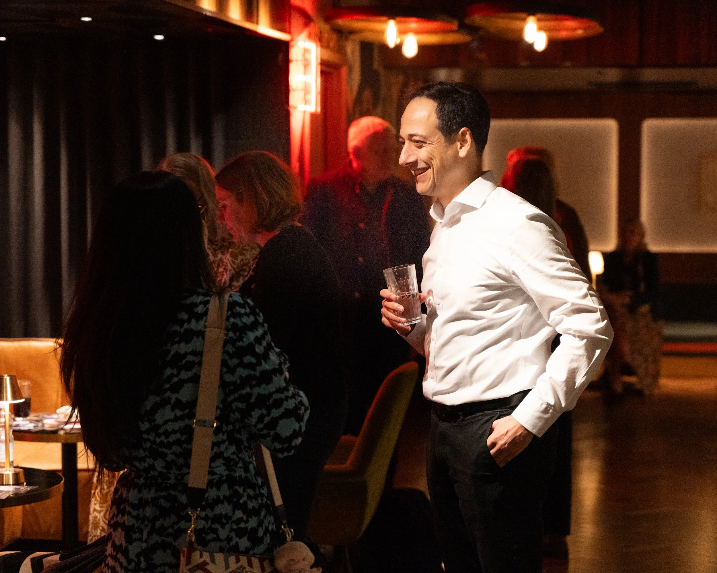 A man wearing a white dress shirt, dressed in black trousers, holding a glass of beverage, standing and smiling indoors with warm lighting, surrounded by people in conversation, and a dark wood-paneled wall in the background.