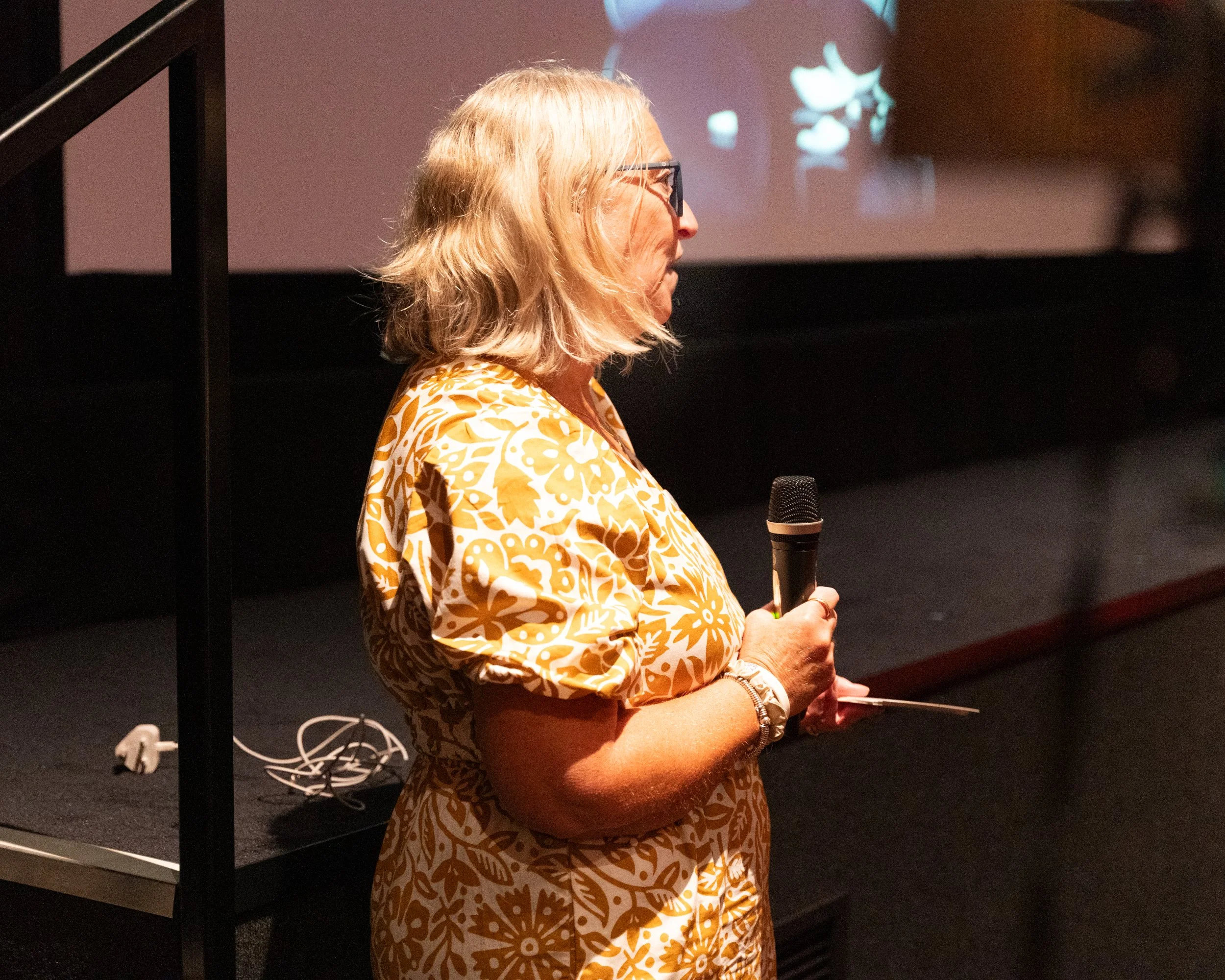 A woman with blonde hair and glasses wearing a yellow and white patterned dress is standing indoors, holding a microphone, and appears to be speaking or presenting.