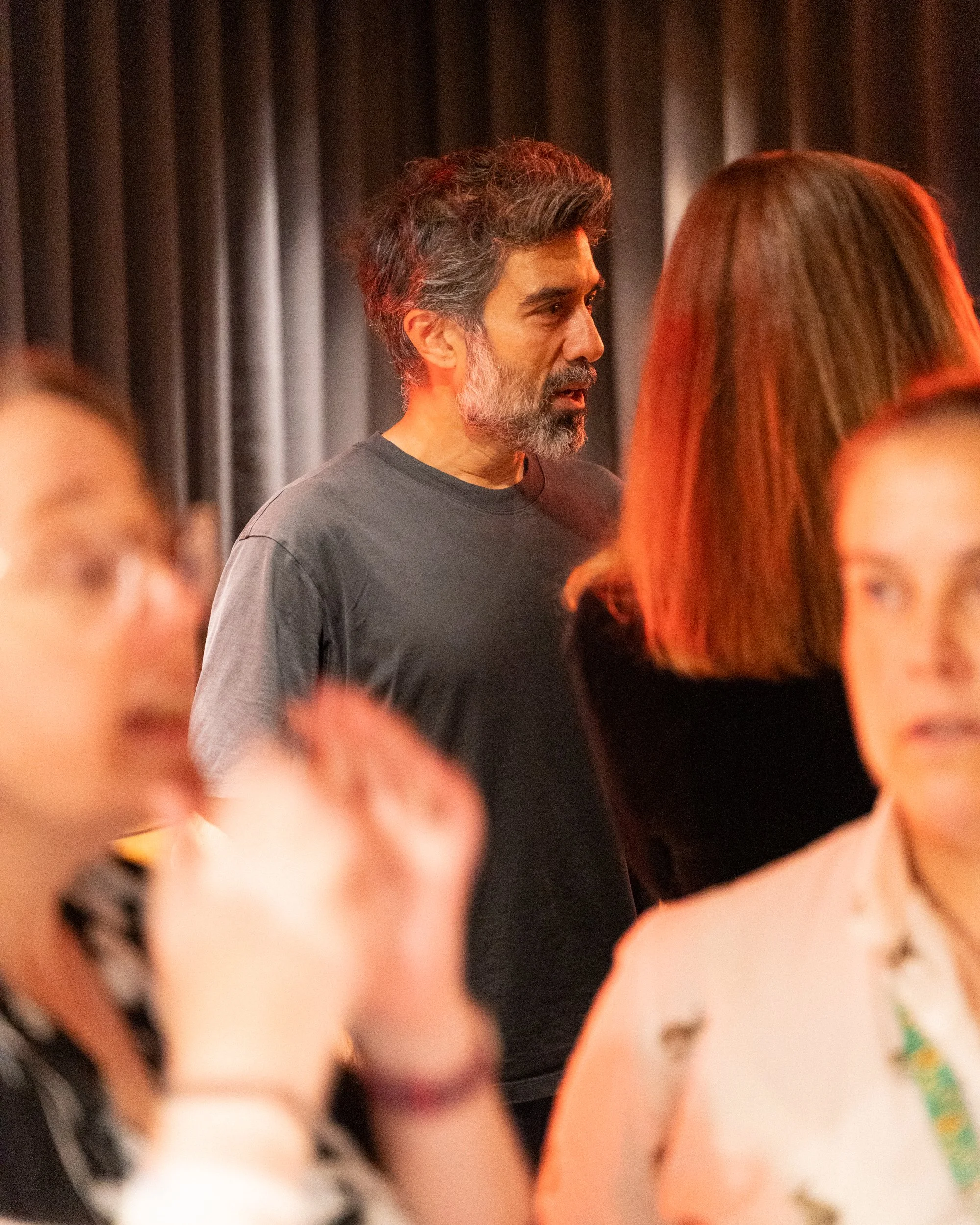 A middle-aged man with salt-and-pepper hair and a beard, wearing a dark T-shirt, appears to be engaged in conversation in a setting with a dark curtain in the background. The foreground shows the blurred heads of two women, one with reddish-brown hai