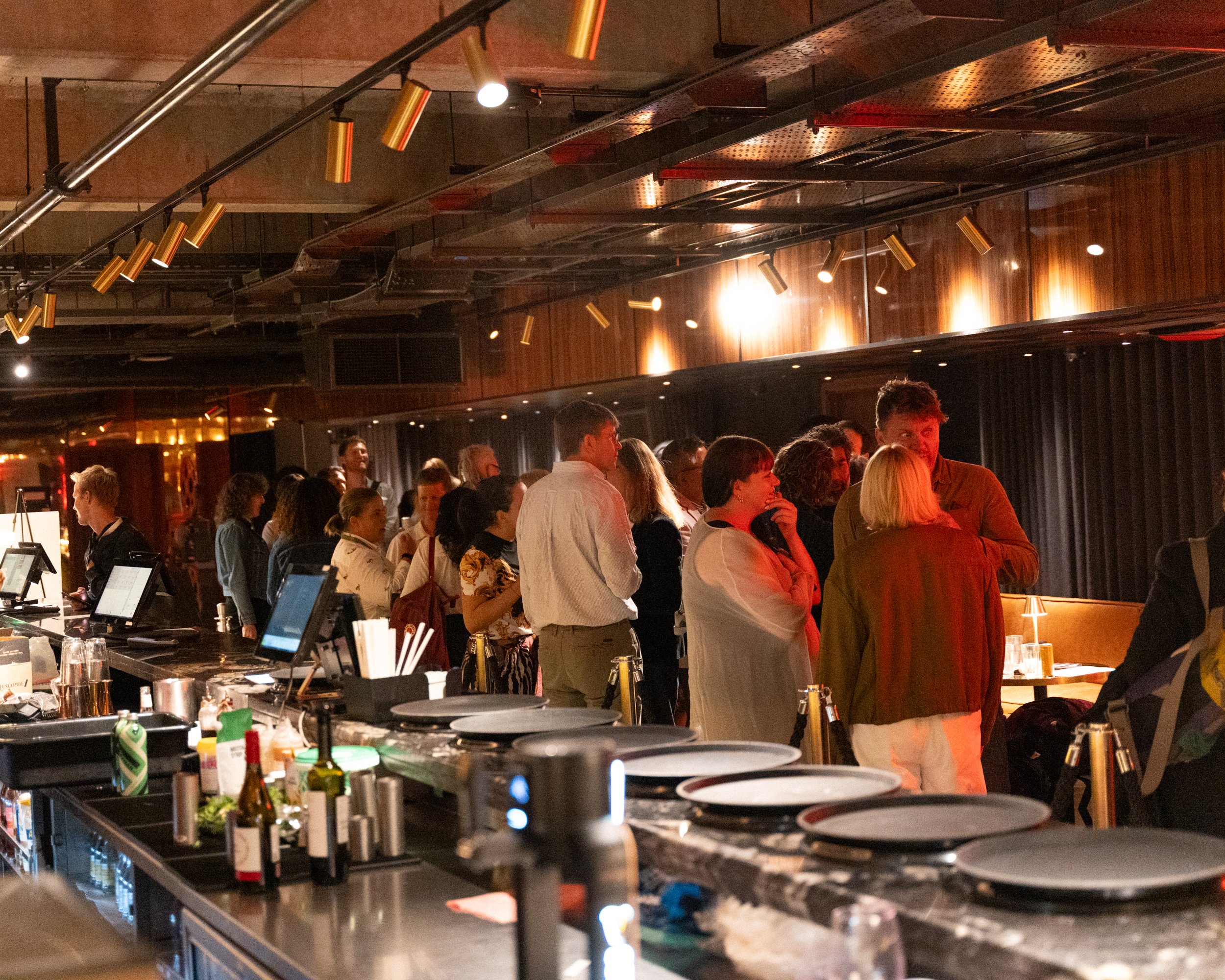 People gathered in a dimly lit bar or restaurant with a modern industrial design, featuring a wood-paneled wall and a ceiling with exposed metal ductwork, overhead lighting, and a bar counter with drinks and utensils.