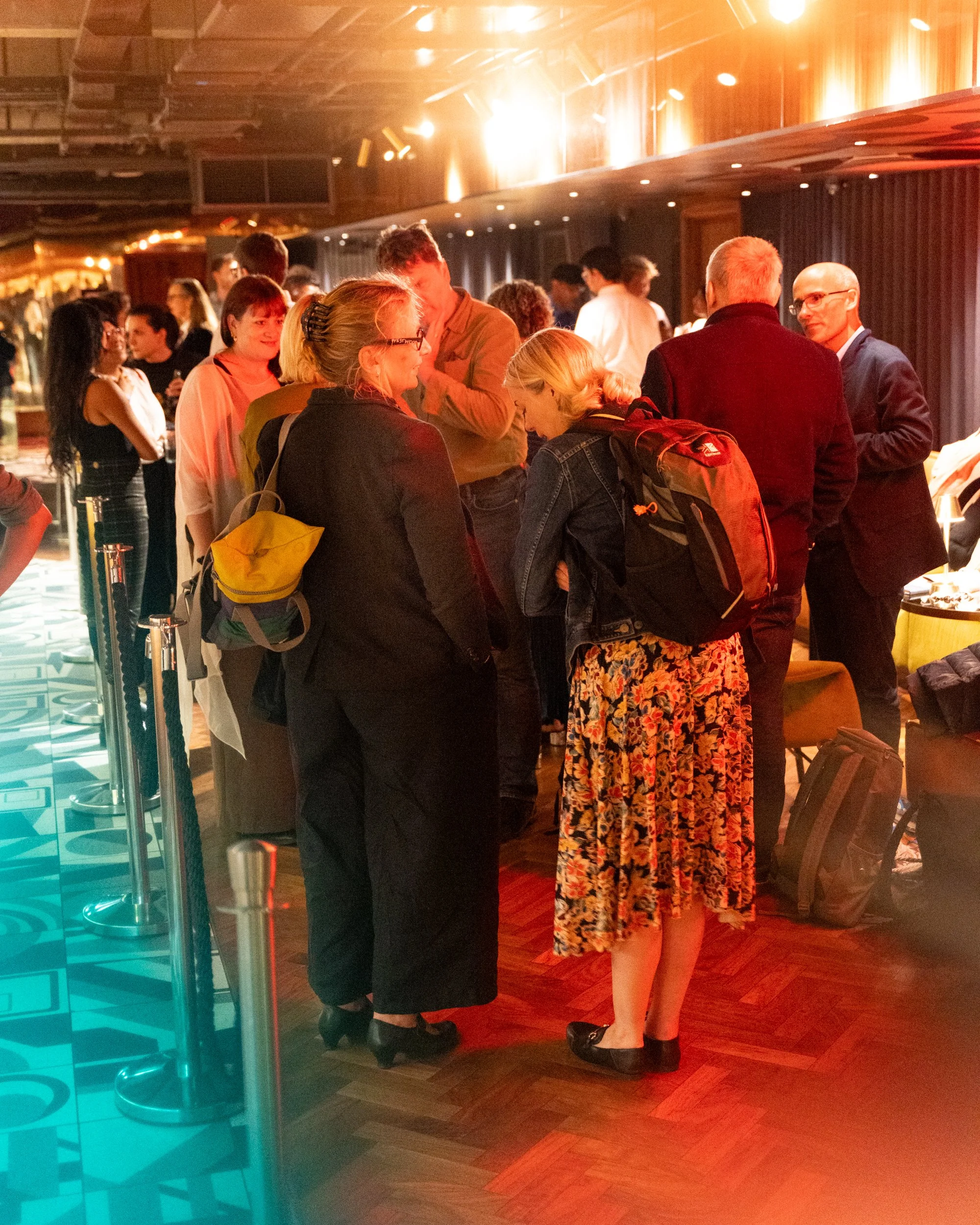 People engaged in conversation in a lively indoor setting, with warm lighting and wooden flooring.