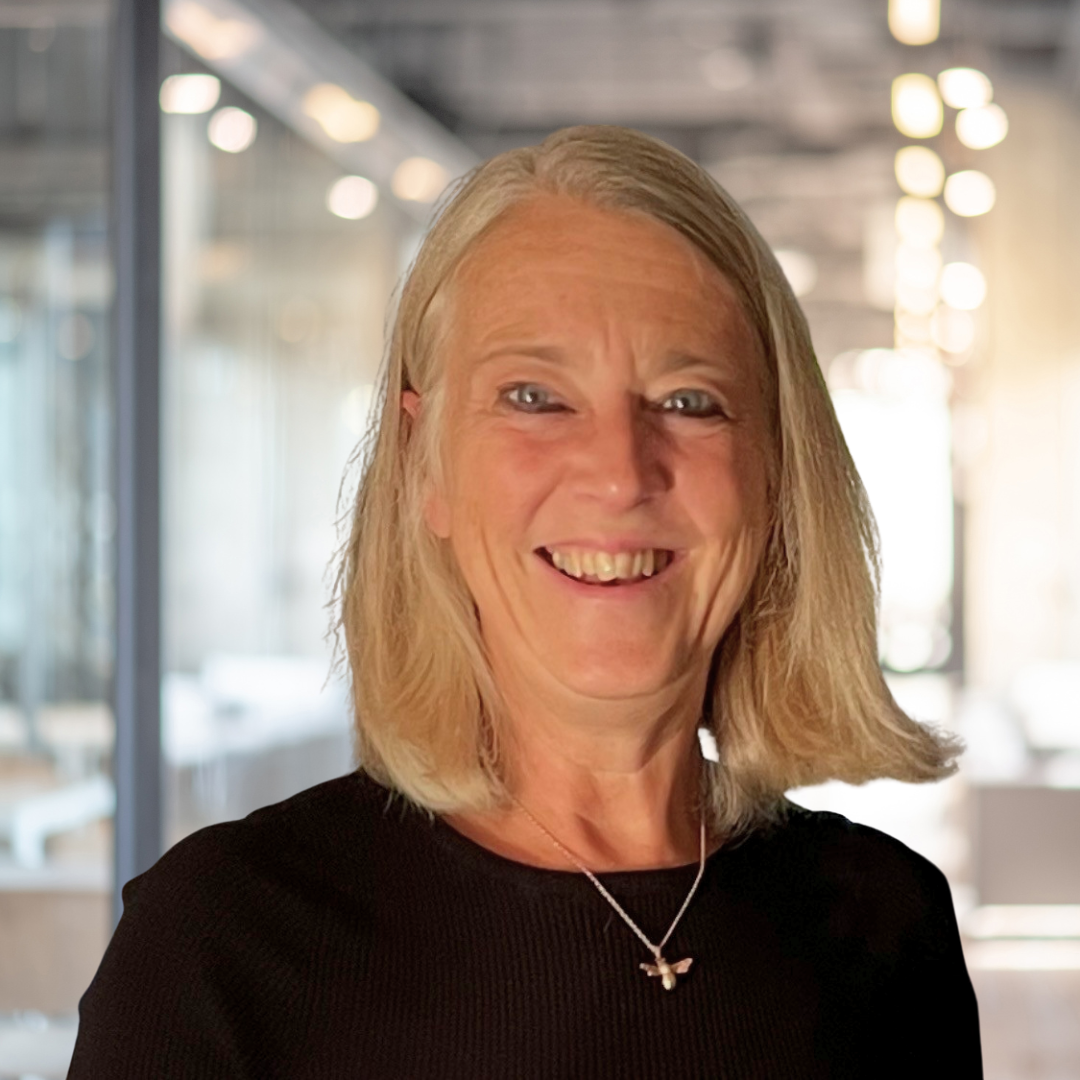A smiling woman with shoulder-length gray hair, wearing a black top and a butterfly necklace, standing in a modern, well-lit indoor space with large windows and blurred background lights.