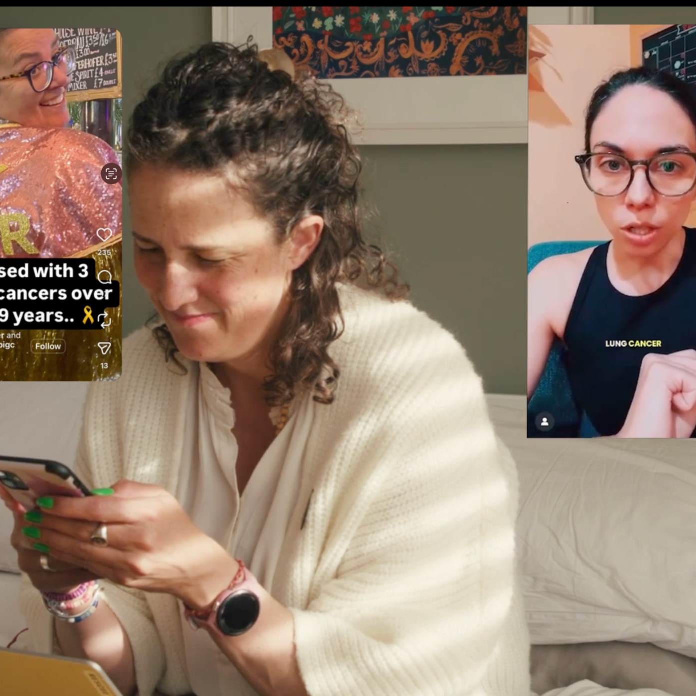 A woman with curly hair and a beige sweater looking at her phone, sitting on a bed, with two large screens behind her showing women talking about health issues, one with text mentioning cancer.