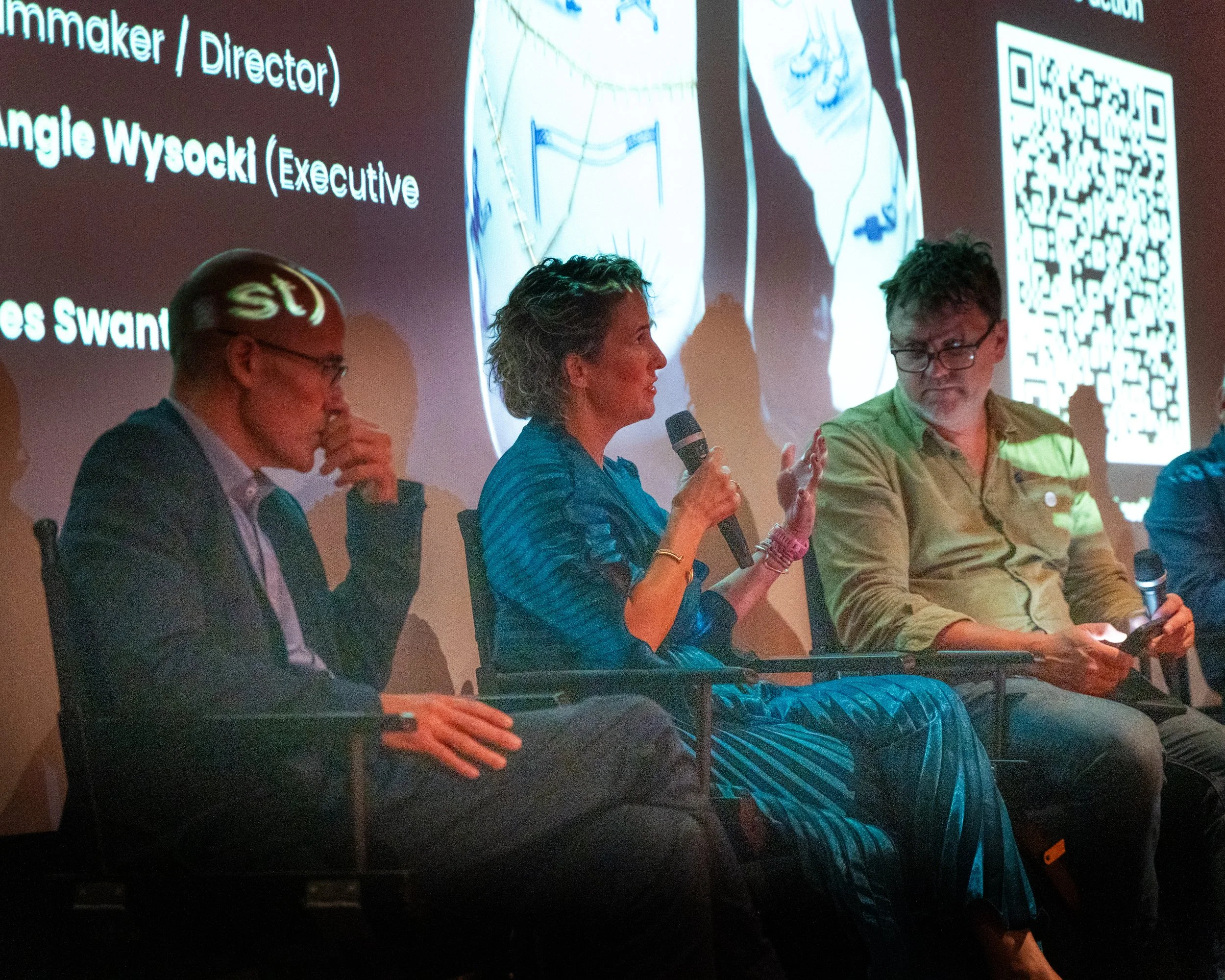 People participating in a panel discussion, two men and a woman, seated and speaking. The woman is holding a microphone, and there's a large presentation screen behind them with text and QR code.