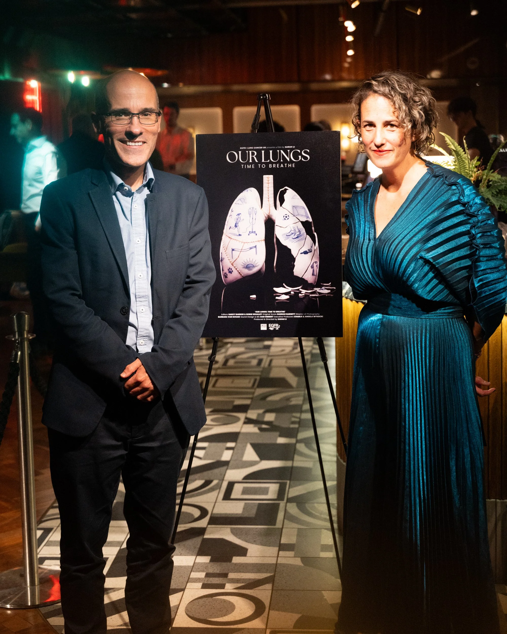 Two people, a man and a woman, standing next to a poster titled 'Our Lungs: Time to Breathe' in a dimly lit indoor setting with a patterned black and white floor. The man is smiling, wearing glasses and a dark suit with a light-colored shirt, while t