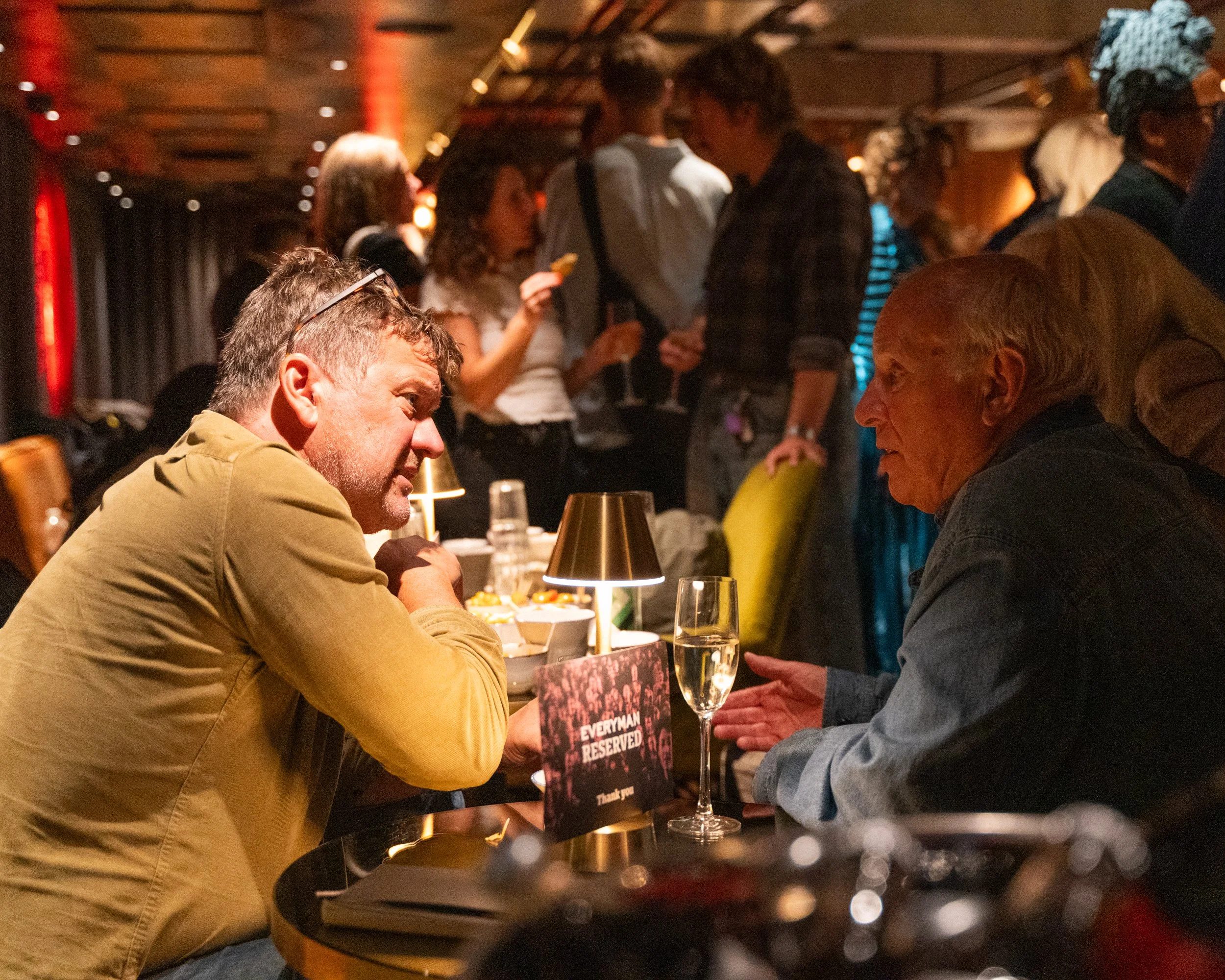 People engaging in conversation at a social event in a dimly lit room, with a table that has a sign reading 'Everyone Reserved' and a glass of champagne in the foreground.
