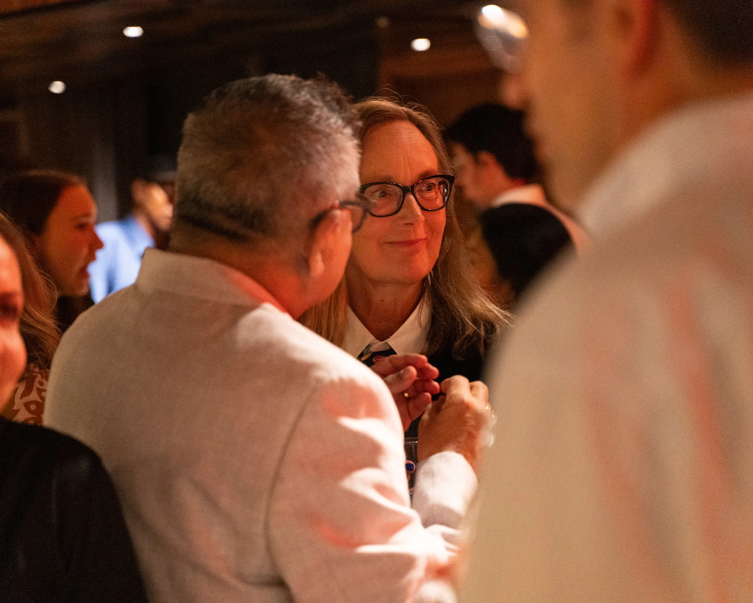 People attending an event, woman wearing glasses smiling, man with gray hair, other guests visible in background, indoor setting with warm lighting.