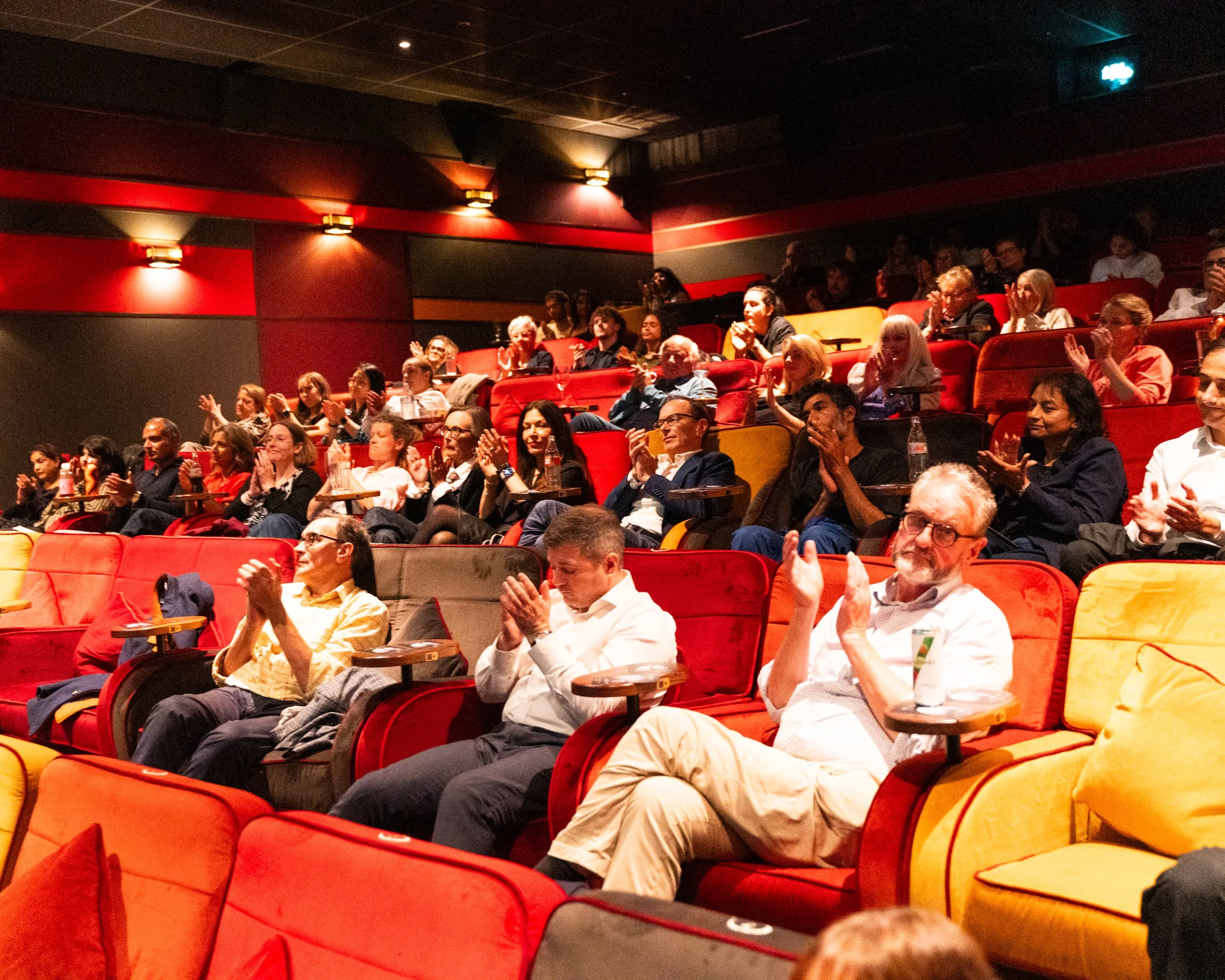 Audience of adults sitting in red and yellow theater seats, clapping and watching a performance in a darkened theater with subdued lighting.