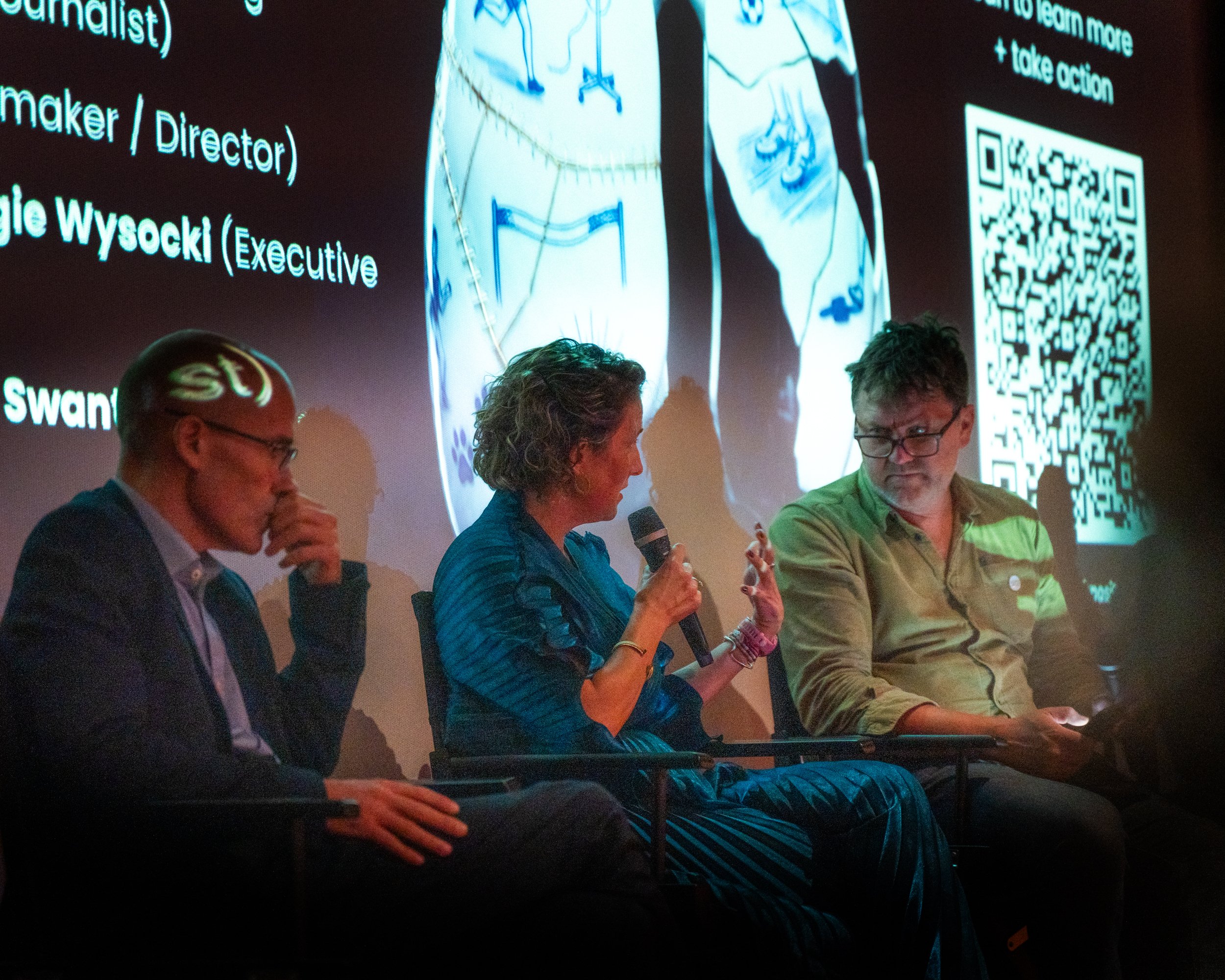 A panel of four people sitting on stage during a discussion, with a large screen behind them displaying text, images, and a QR code. The audience appears to be in a dark room.