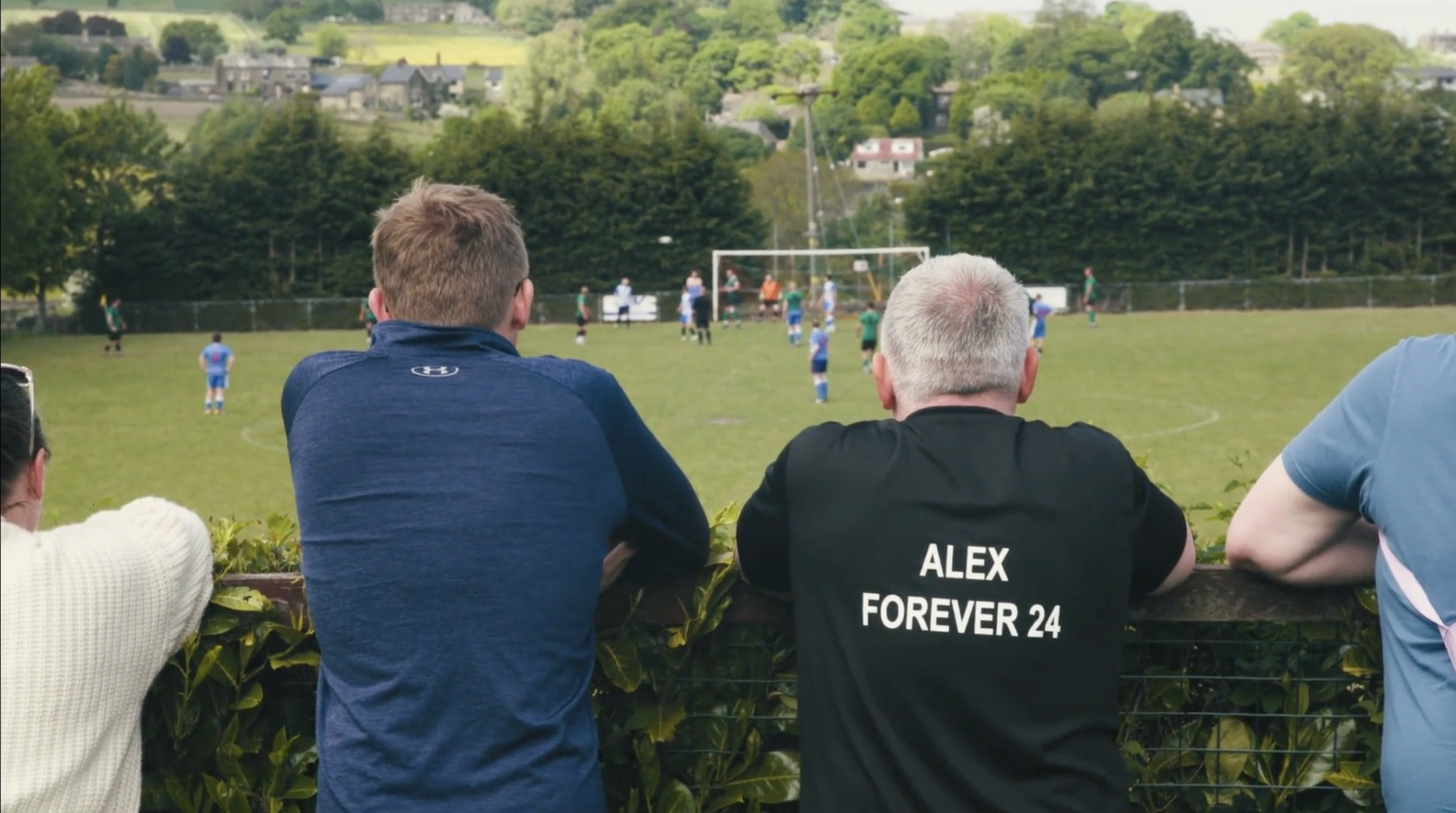 Spectators watching a soccer game on a grassy field with trees and houses in the background, one person wearing a black shirt with the name "ALEX FOREVER 24."